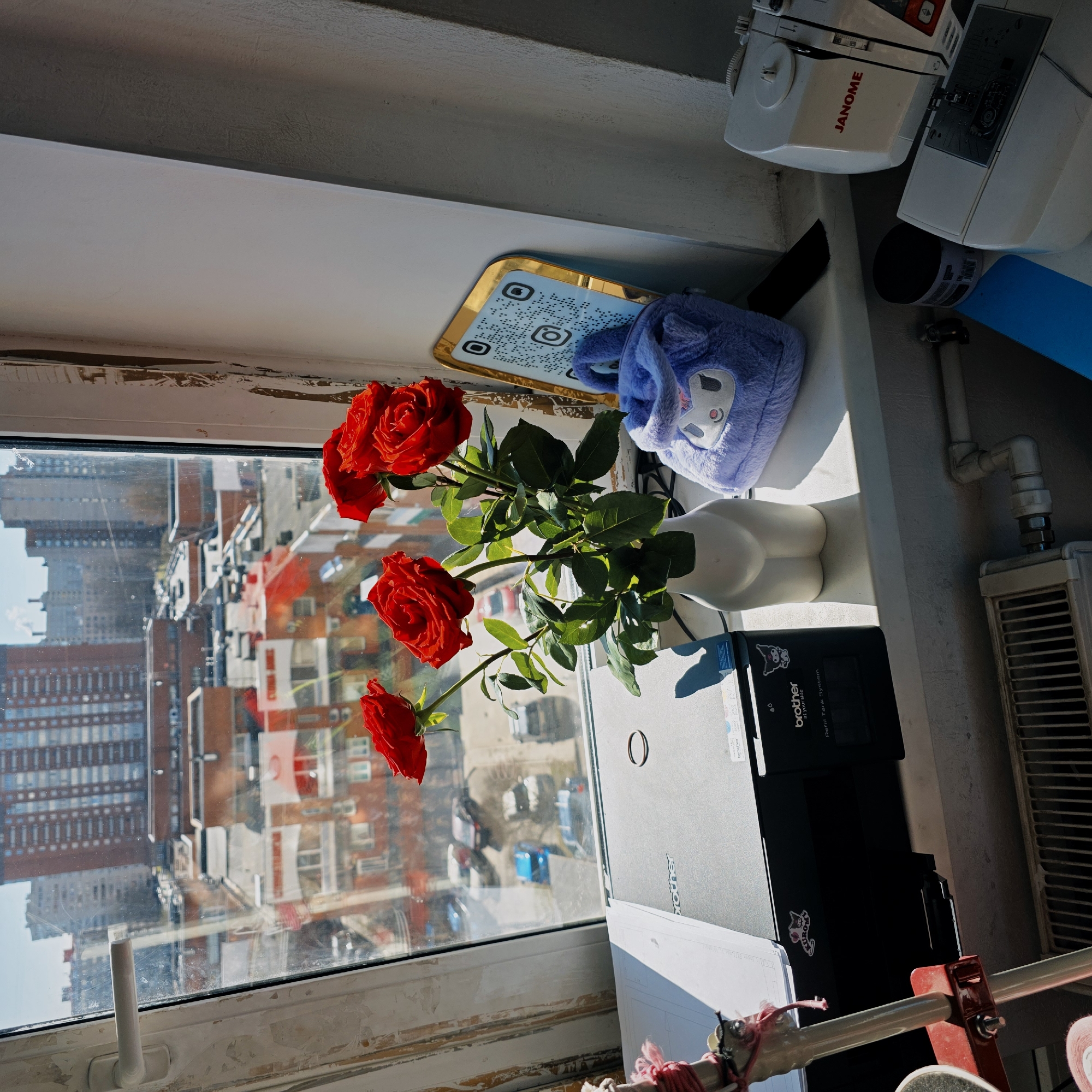 A windowsill scene with a view of Saint Petersburg, Russia. Three red roses in a white vase are positioned in front of a window. A QR code on a gold tray and a blue fuzzy bunny are placed nearby. A Janome sewing machine, a black Brother printer, a blue mat, and a radiator are also located on the windowsill. The view outside the window includes numerous brick buildings. The window frame is white but appears aged.