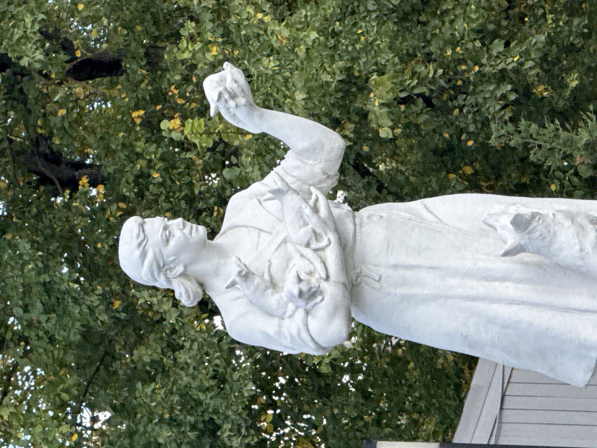 A white stone statue depicts a woman holding several small animals in her arms and hand. The woman, with her hair tied back, wears a simple dress and stands outdoors against a backdrop of lush green trees, some with yellowing leaves, indicating autumn. She is looking to her right. In her left arm, she cradles at least three small animals, possibly puppies or kittens, which appear to be nursing or nestled together. In her outstretched right hand, she holds another small animal. At the base of the statue, a small animal, similar to those she is holding, is visible. The statue is situated in a park-like setting, with a section of a light-colored wooden fence visible in the lower-left corner. The statue is located in Novodevichy Cemetery, Moscow, Russia.