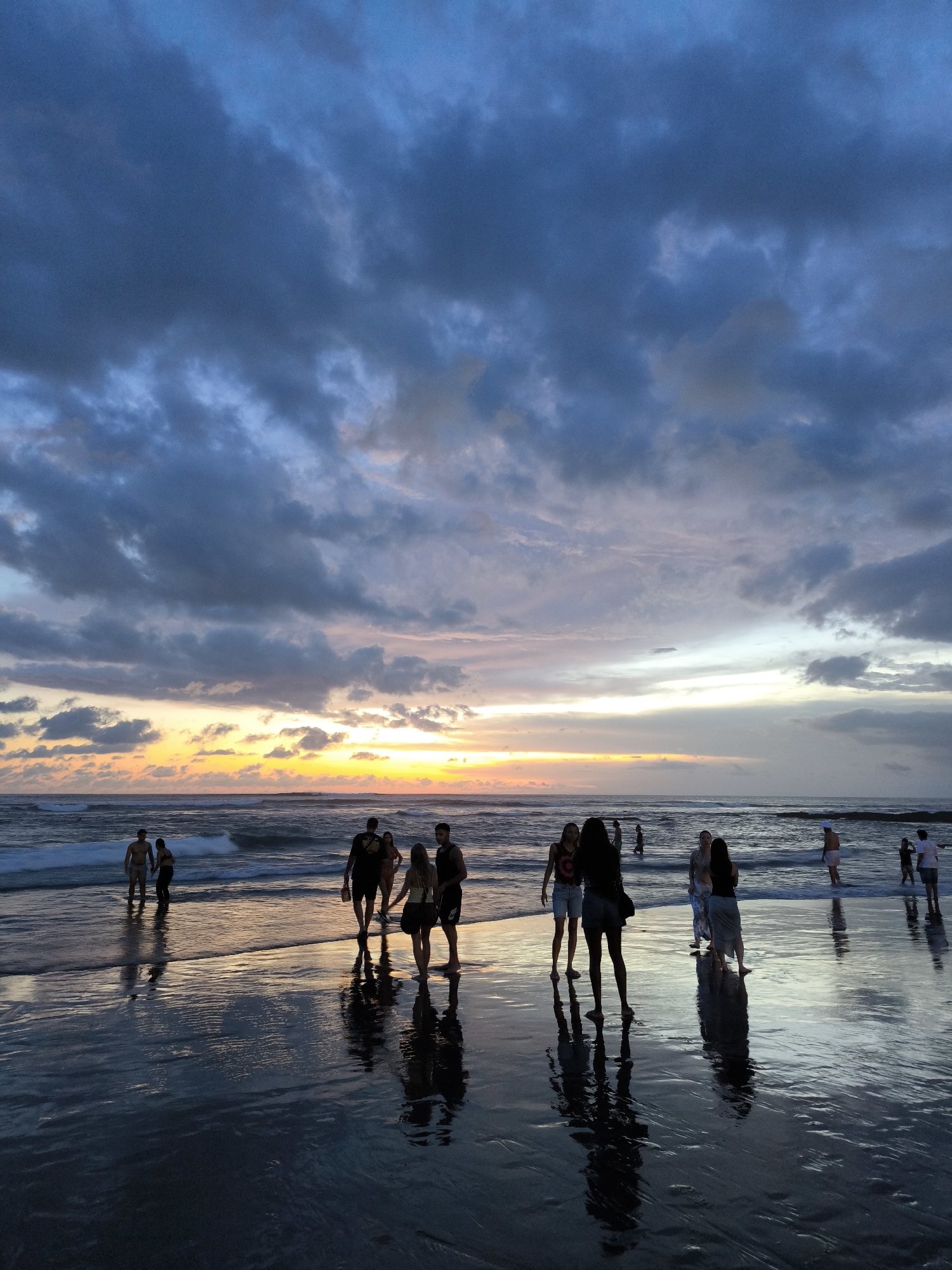 The image depicts a beach scene at dusk in Canggu, Indonesia. A group of people are scattered across the wet sand of the beach, many wading in the shallow water. Their figures appear as silhouettes against the colorful horizon where the sun is setting, creating a gradient of yellow, orange, and purple in the sky. The sky is also covered with dark, heavy clouds. The wet sand reflects the colors of the sky, doubling the visual impact of the sunset. Waves are gently rolling onto the shore. The people are interacting, some walking together, others standing and observing the sunset.