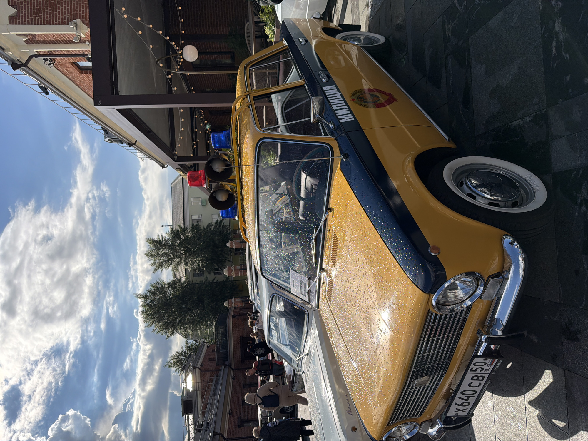 The image depicts a yellow and blue classic car parked on a street in Moscow, Russia. The car has the word "МИЛИЦИЯ" (Militia) written on its side, an old police force name in Russia. It's equipped with vintage-style red and blue lights on top. Water droplets are visible on the car's surface, indicating recent rain. People are walking in the background, suggesting a public area. The backdrop includes buildings and trees, with a cloudy sky above. A license plate is partially visible on the front of the car.