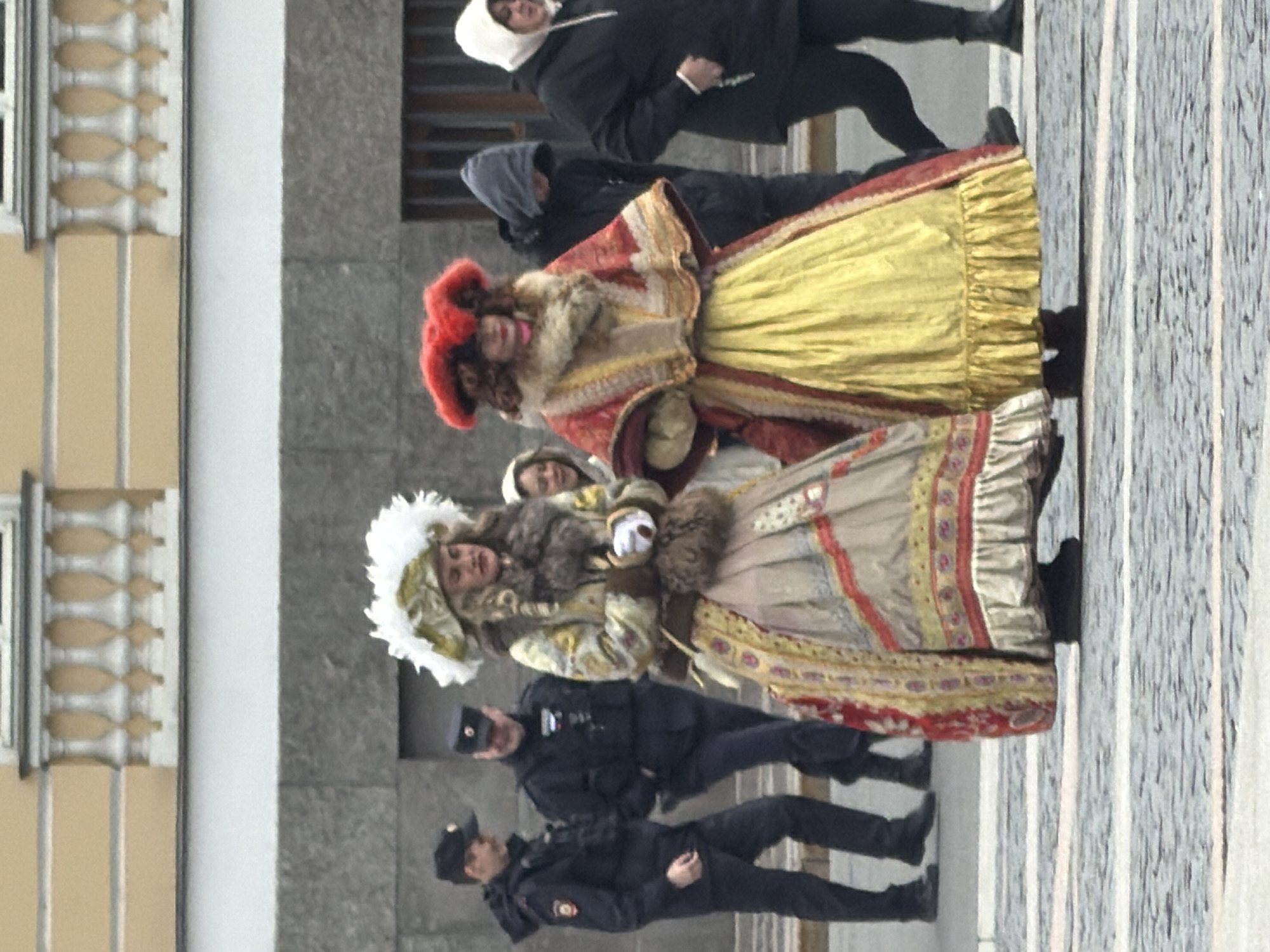 The image captures a street scene in Saint Petersburg, Russia. Two women dressed in ornate, historical costumes are the focal point. Both wear elaborate gowns with fur accents and large, decorative headpieces. Behind them, two men in dark hooded jackets are visible. In the foreground, two uniformed police officers stand in front of the women in costumes. The background shows an architectural facade with distinct window moldings. The street surface consists of light gray planks.