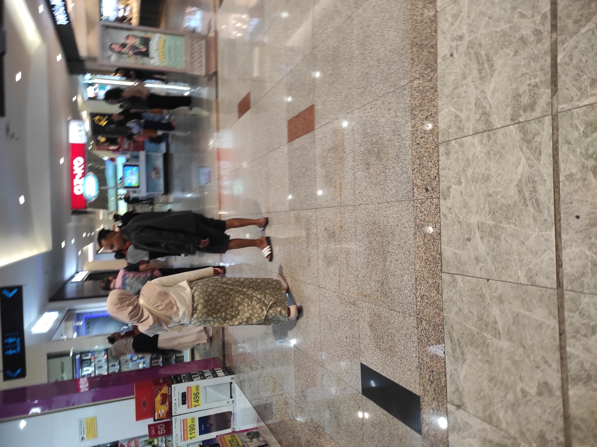 The image depicts the interior of a shopping mall in Surabaya, Indonesia. In the foreground, two individuals are walking away from the camera on a polished, speckled tile floor. The person on the left is wearing a beige hijab, a white long-sleeved top, and a long, patterned green skirt. The person on the right is wearing dark shorts and a black t-shirt, with black and white flip-flops. Their bodies are angled away from the viewer.
In the midground, several other people are visible, some browsing in shops and others walking. The shops are brightly lit with various signage, including a red sign that says "OZ.KO". To the left, there are displays of products, some featuring prices like "HARGA SPESIAL 1.190.000" and "1.496.000". Further back, a directional sign with arrows and the word "TOILETS" is mounted on the ceiling. The environment is spacious, with high ceilings and fluorescent lighting. Reflections of the lights and surroundings are visible on the floor. The overall scene suggests a typical commercial area within a mall.