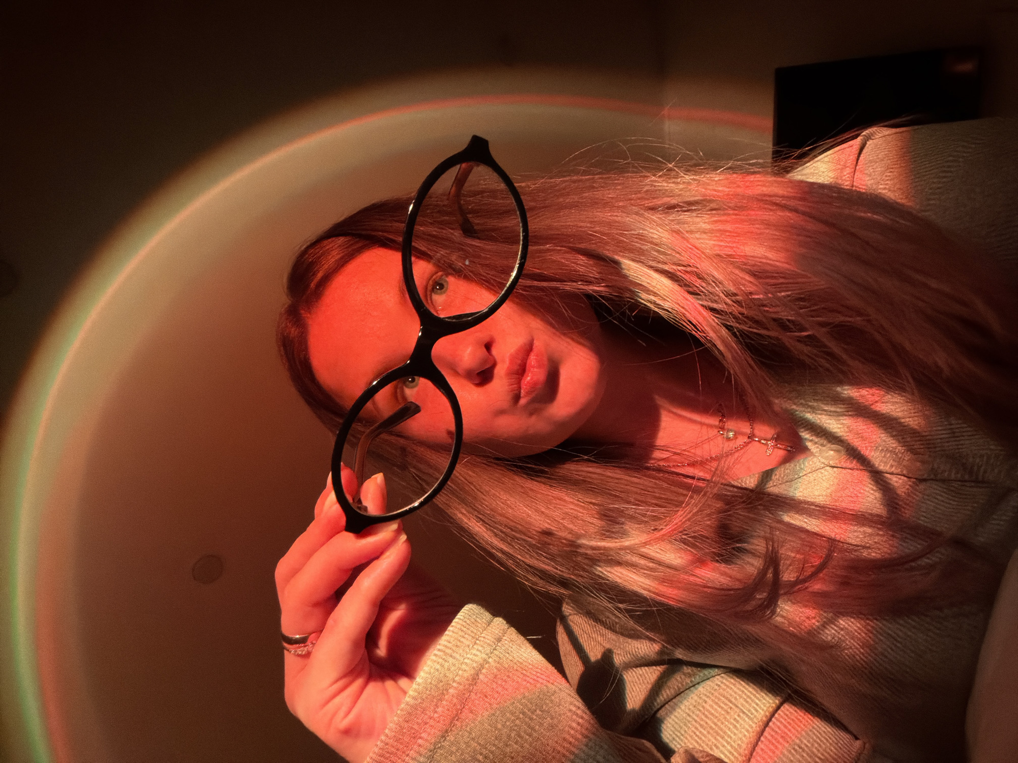 A person with long blonde hair is holding up a pair of black glasses, partially obscuring their face. The lighting is predominantly warm orange, with a rainbow-like color effect visible in the background, suggesting an artificial light source or filter. The person is wearing a light-colored patterned shirt. The background is blurred and features a light wall and a dark rectangular object. The image was most likely taken in an indoor setting, though the precise location within the city of Chaikovsky, Russia cannot be determined from the image.