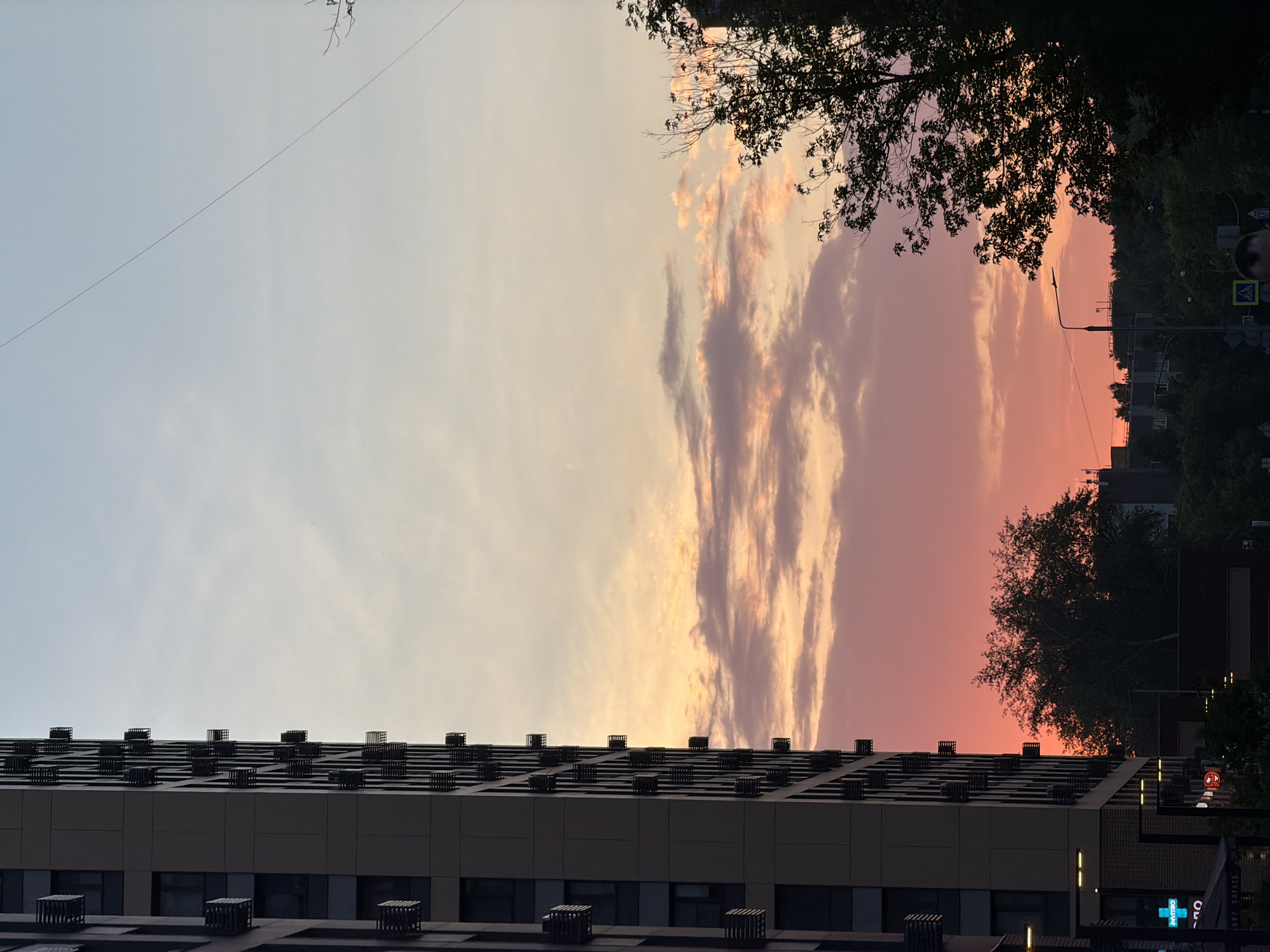 The image captures a city skyline during sunset, likely in Moscow, Russia. A tall, modern apartment building dominates the left side of the frame, its dark facade contrasting with the vibrant sky. The building features numerous balconies or external units, arranged in a repeating pattern. To the right, silhouetted trees and other buildings form a darker, lower silhouette against the colorful clouds. The sky transitions from a pale blue at the top to shades of orange, pink, and red near the horizon, indicating the sun is setting. A thin electrical wire stretches diagonally across the upper portion of the sky. In the foreground, street-level details such as a building with illuminated signs and part of a street with a traffic light and pedestrian crossing are visible. A person's head is partially visible at the bottom center of the image. The scene appears to be a typical urban environment experiencing the end of the day.