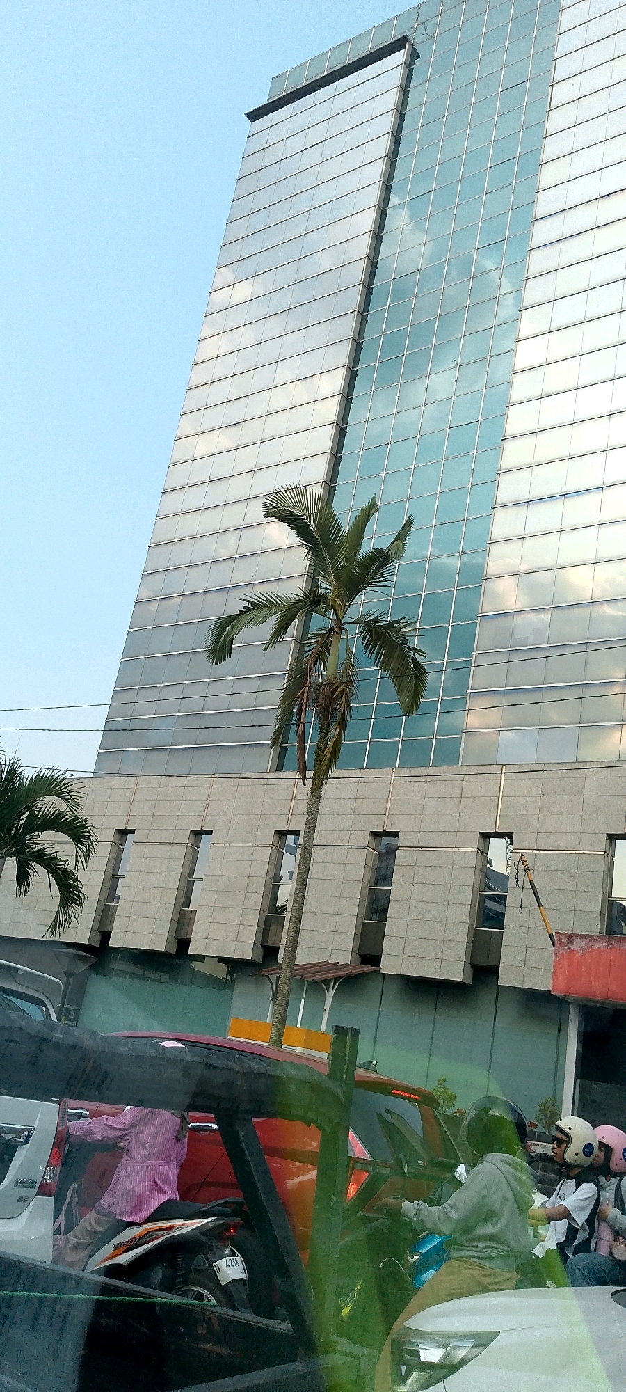 In Bandung City, Indonesia, a street scene unfolds beneath a towering glass-fronted building. The skyscraper's facade is composed of numerous rectangular windows, reflecting the sky and clouds. In the foreground, a slender palm tree with sparse fronds stands tall, its trunk partially obscured by the building's lower structure, which features a series of slanted concrete panels with rectangular windows.
The lower part of the image depicts a traffic jam. Several vehicles, including cars and motorcycles, are visible. A person wearing a pink striped shirt and light-colored pants is seen reaching out from a white car. On a motorcycle, a person in a grey hoodie and light brown pants is seated, facing forward. Behind them, two other individuals are on a motorcycle, one wearing a white helmet with black stripes and the other a pink helmet. The overall scene suggests a typical urban environment with heavy traffic and modern architecture.