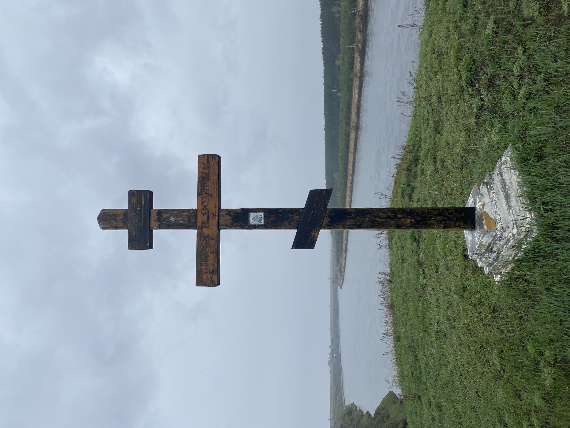 A wooden Russian Orthodox cross stands on a grassy bank overlooking a body of water, possibly a river, under an overcast sky. The cross is dark-stained and has the characteristic three crossbars of the Russian Orthodox tradition. There is a small, faded icon affixed to the center of the cross. The base of the cross is supported by a painted concrete or stone foundation. The location is identified as Novomaltinsk, Russia.