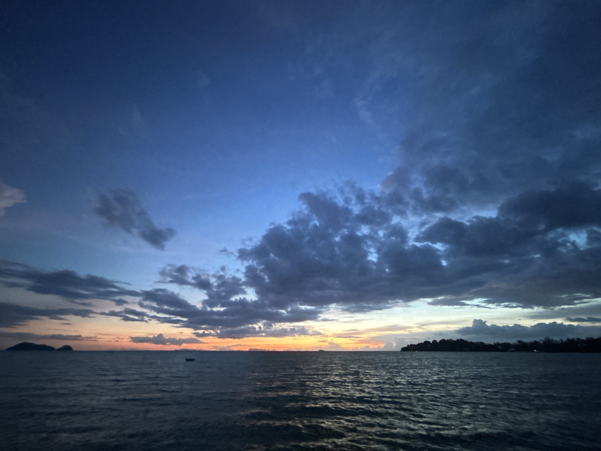This is a wide-angle, eye-level photograph of the ocean at dusk, taken from Ko Pha-ngan District, Thailand. The sky is a deep blue, with scattered clouds, some of which are tinged with orange and yellow from the setting sun. The ocean is a dark, choppy expanse, reflecting the colors of the sky. In the distance, islands and a faint coastline are visible. A small, dark boat is situated on the water, to the right of the center. The overall impression is one of a peaceful, serene twilight scene.