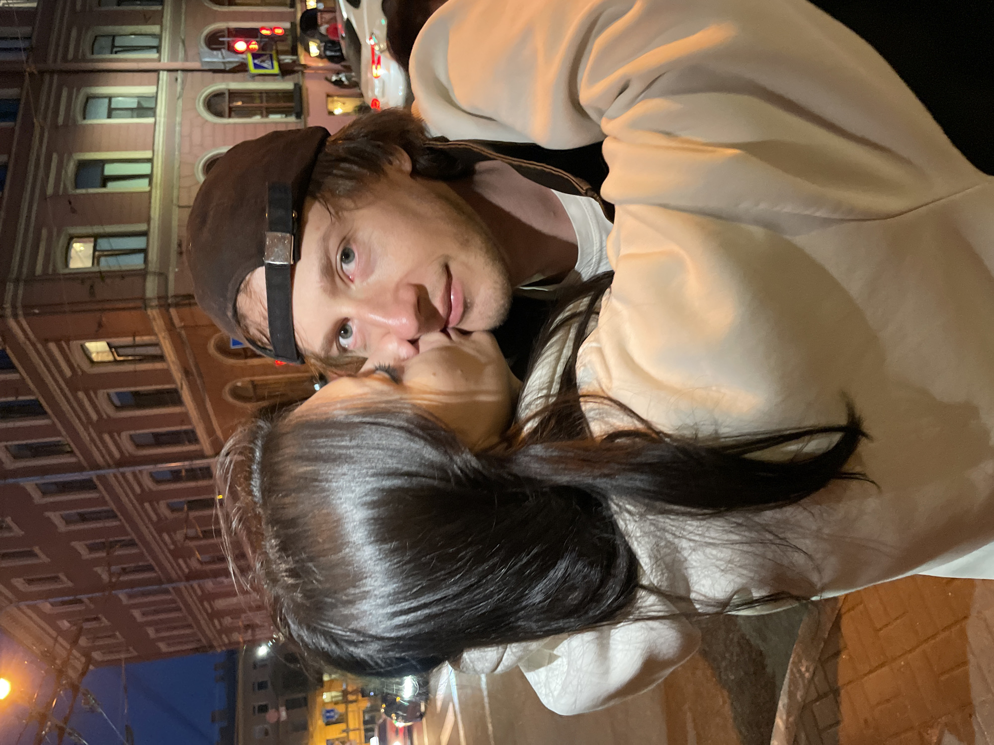 This is a medium shot, taken at night in Saint Petersburg, Russia, featuring a couple in an intimate embrace. A woman, partially obscuring her face with her dark hair, is kissing a man on the cheek. The man, wearing a dark baseball cap and a light-colored hoodie over a white t-shirt, looks towards the camera with a neutral expression. They are standing on a brick-paved sidewalk next to a street with traffic lights and vehicles visible in the background. Behind them, a red brick building with multiple windows and architectural detailing is illuminated by streetlights. The overall atmosphere suggests a romantic moment captured during an evening outing in the city.