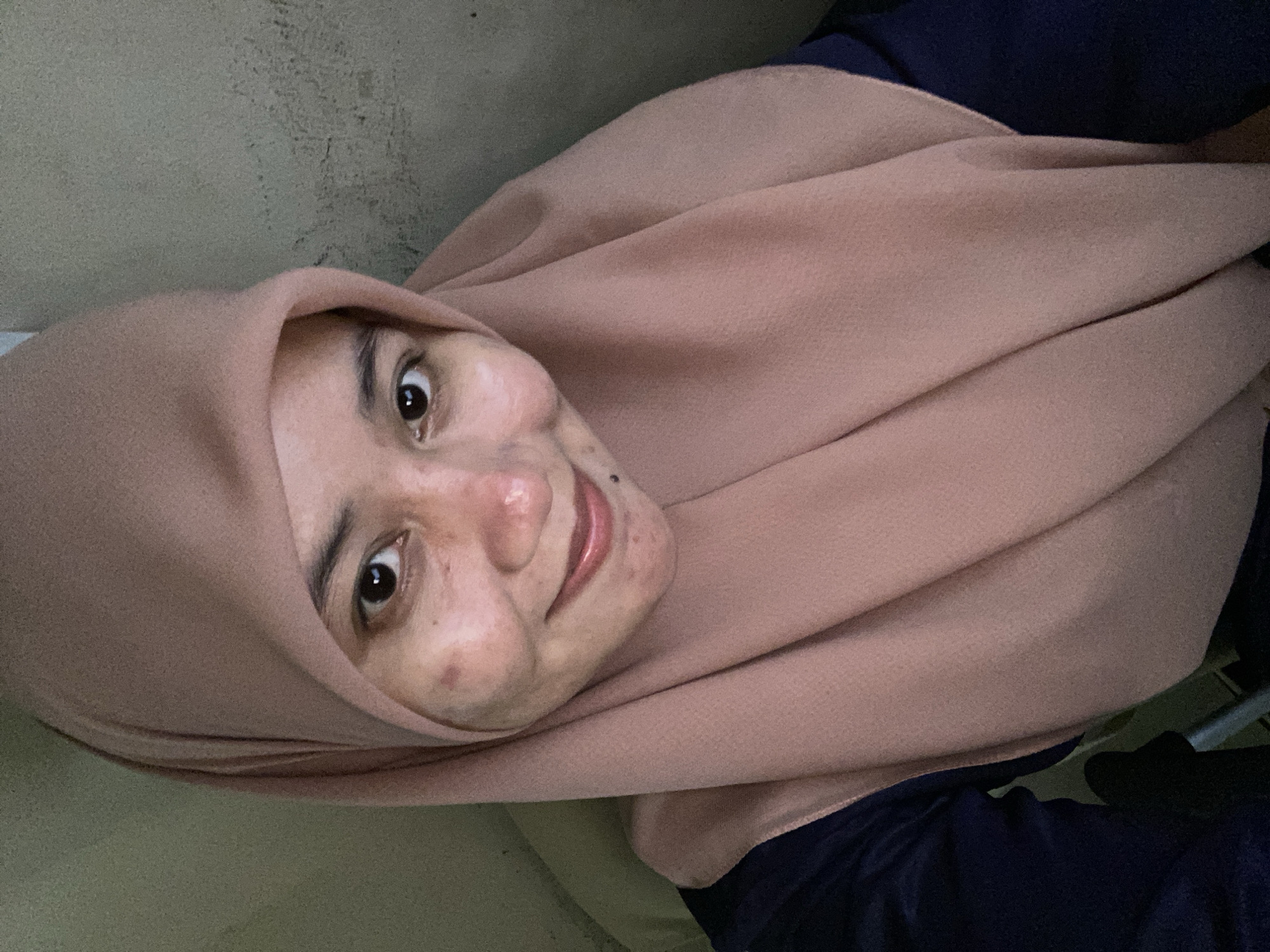 A selfie is taken from a slightly high angle. A woman wearing a light pink hijab and a dark blue top is the subject. Her face is turned slightly towards the camera, and she is smiling. She has visible acne scars and blemishes on her cheeks and forehead. The background is a plain, textured wall, appearing to be indoors. The lighting is soft and somewhat dim. This image was captured in Banjarbaru, Indonesia.