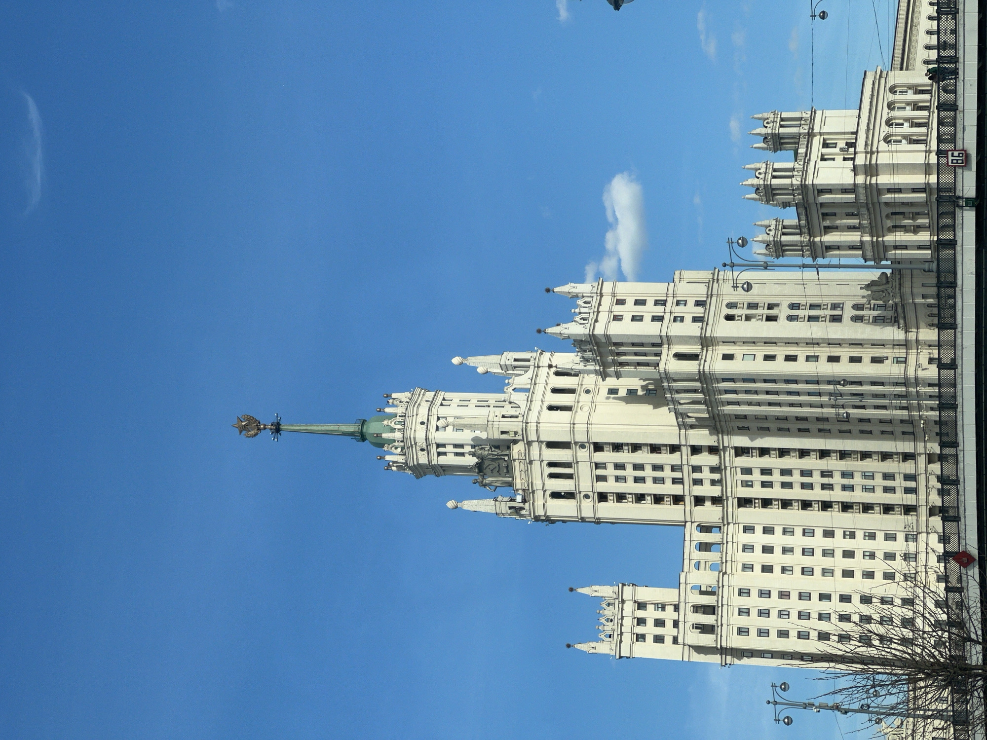 The image depicts the Kotelnicheskaya Embankment Building, one of the Seven Sisters skyscrapers in Moscow, Russia. The tall, white building dominates the frame with its elaborate Stalinist architecture, including multiple tiers, spires, and decorative elements. The clear blue sky is visible in the background. Bare tree branches are at the bottom right of the image. A bridge and overhead wires appear on the right side.