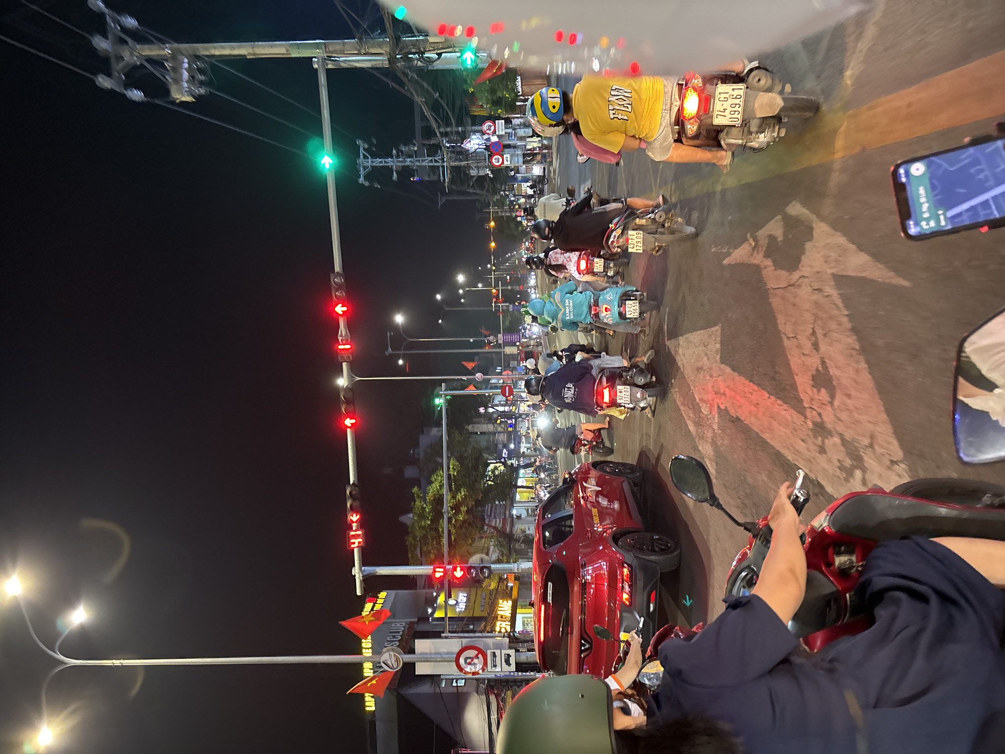 This image captures a nighttime scene on a busy street in Đà Nẵng, Vietnam. The street is filled with motorbikes waiting at an intersection, facing a traffic light that shows red. Several motorbikes are visible at the front of the line, waiting for the light to change. A red car is also stopped in the intersection. The person taking the photo is also on a motorbike. Streetlights illuminate the scene. Buildings and street signs are visible along the side of the road, including a sign for S Club.