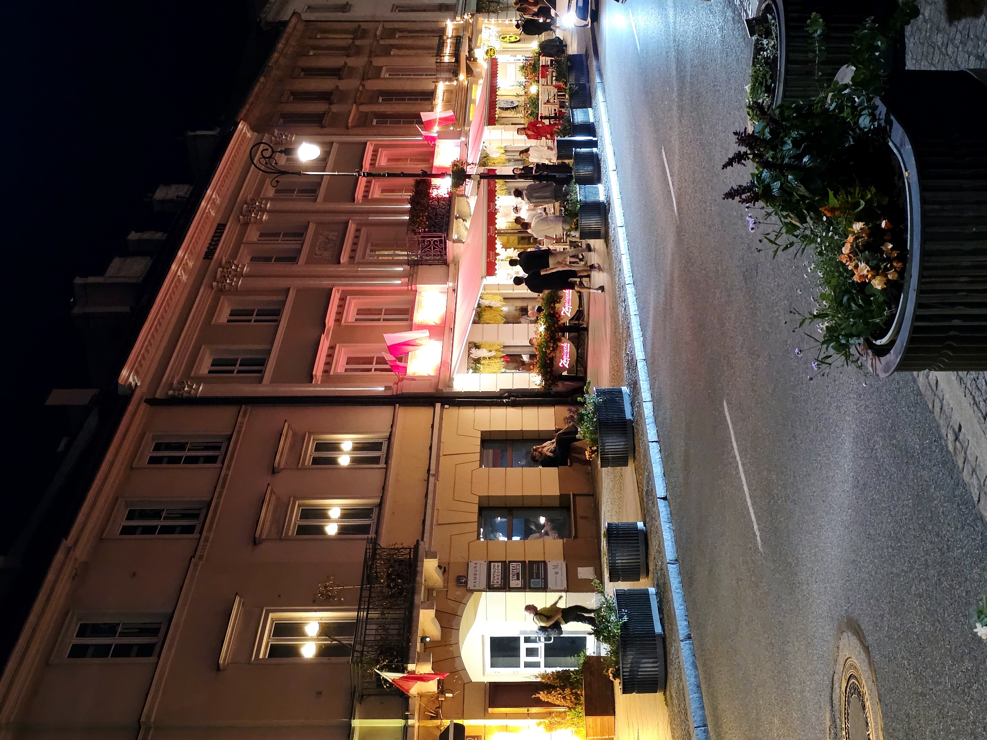 This is a nighttime photograph of a street scene in Warsaw, Poland. The image captures a section of a building lining a city street, which appears to be a commercial area with restaurants and shops.
The building is multi-story, with a pale facade and numerous windows. The architecture suggests a historical or classical style. Several windows on the upper floors are illuminated, casting a warm glow. Red awnings are visible over the ground-floor establishments, adding a splash of color to the scene. Polish flags, red and white, are displayed on poles outside some of the windows.
The street itself is paved and has a single white line marking a lane. There are several large, dark-colored planters containing greenery and flowers lining the sidewalk. A streetlamp with a decorative curved pole is visible, providing additional illumination.
Several people are present in the image. In the foreground, on the sidewalk, there are groups of people walking and conversing. Some appear to be patrons of the establishments. A person with a backpack is seen entering a doorway on the left side of the frame. A couple is seated on a bench in front of one of the buildings.
The overall atmosphere is one of a lively urban evening. The lighting from the street, buildings, and possibly car headlights (a portion of a white car is visible on the right) creates a well-lit but also somewhat shadowed environment. The presence of flags and the architectural style hint at a location within Warsaw, likely a historic or central district.