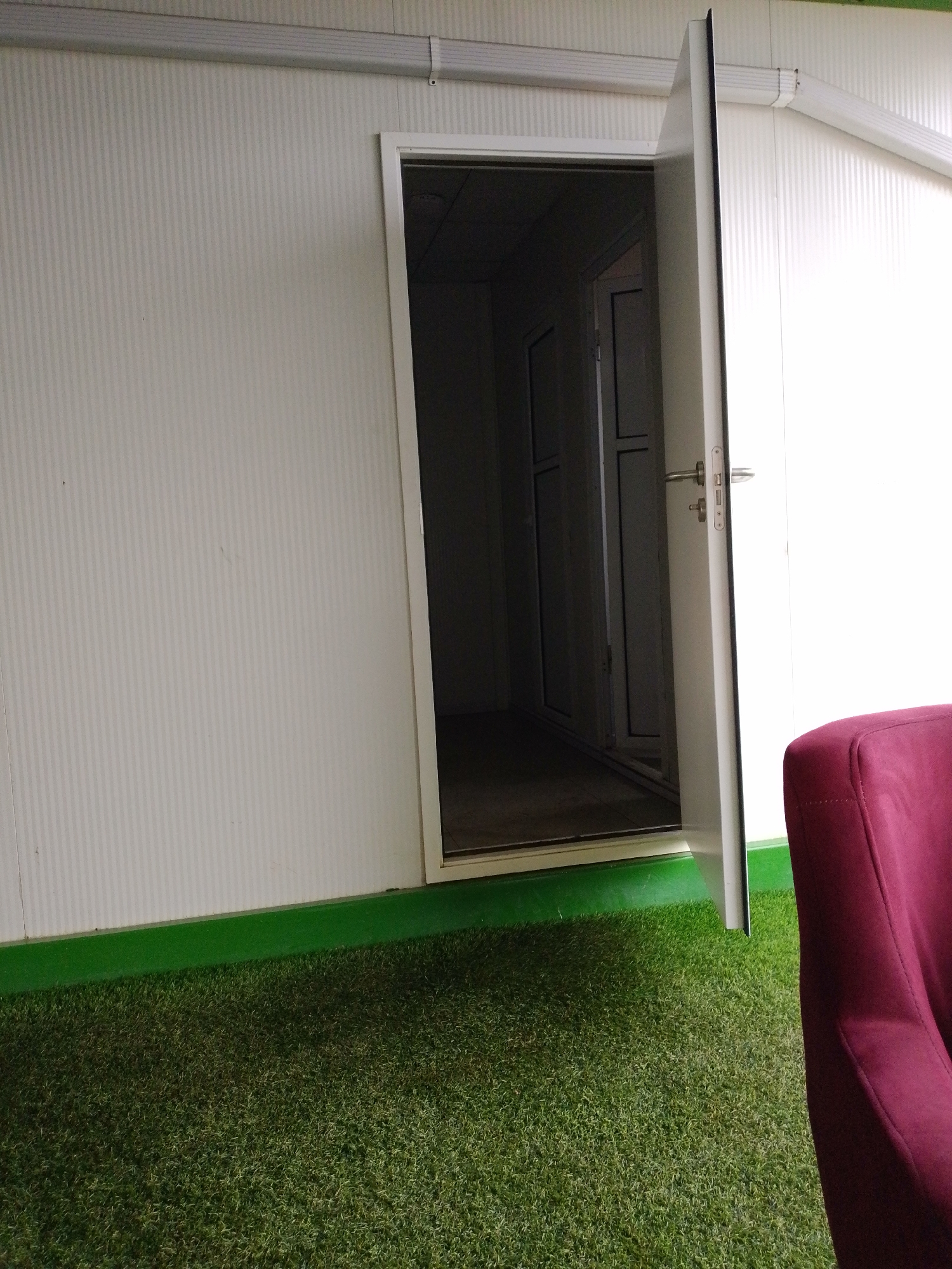 The image shows an open white door revealing a dark interior with another door visible inside. The exterior of the building is white with vertical ribbing. A green strip runs along the base of the wall, transitioning to artificial grass flooring. To the right, a portion of a dark red upholstered chair is visible. The scene appears to be an indoor space, possibly a modular building or room, situated in Barikin Jirga Summa, Nigeria. There are no people visible in the image.
