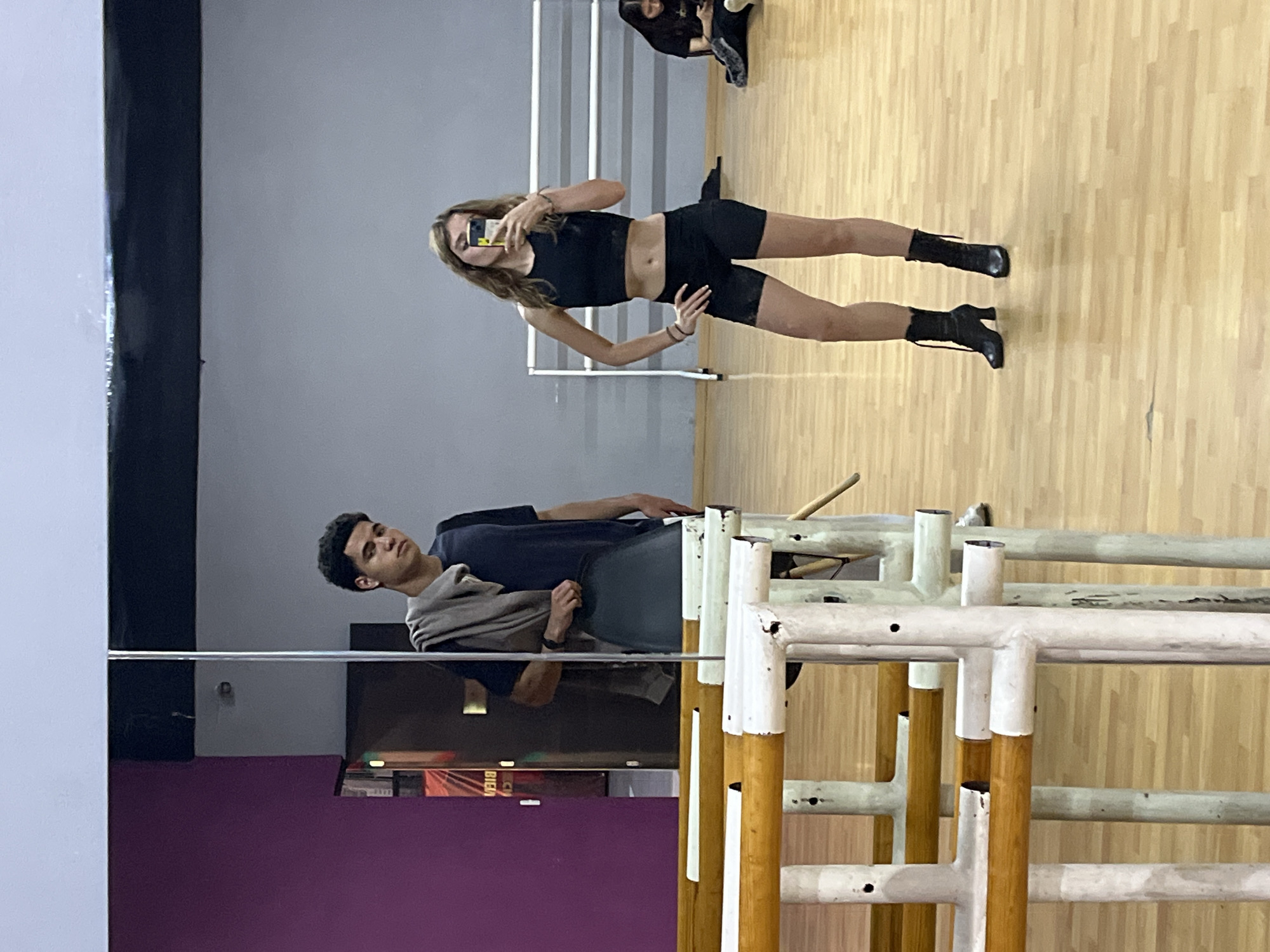 A mirrored dance studio in San Bernardino Tlaxcalancingo, Mexico is shown. A young woman in a black crop top and shorts, wearing black boots, poses and takes a photo of herself in the mirror with her cell phone. Her reflection is captured. A young man wearing a dark t-shirt and a gray shawl stands next to a ballet barre. He is also reflected in the mirror. The background behind the man includes a gray wall, a purple wall, and a poster with Spanish text. The floor is made of light brown wood. In the upper part of the mirror, another person's legs and feet are visible in the background.