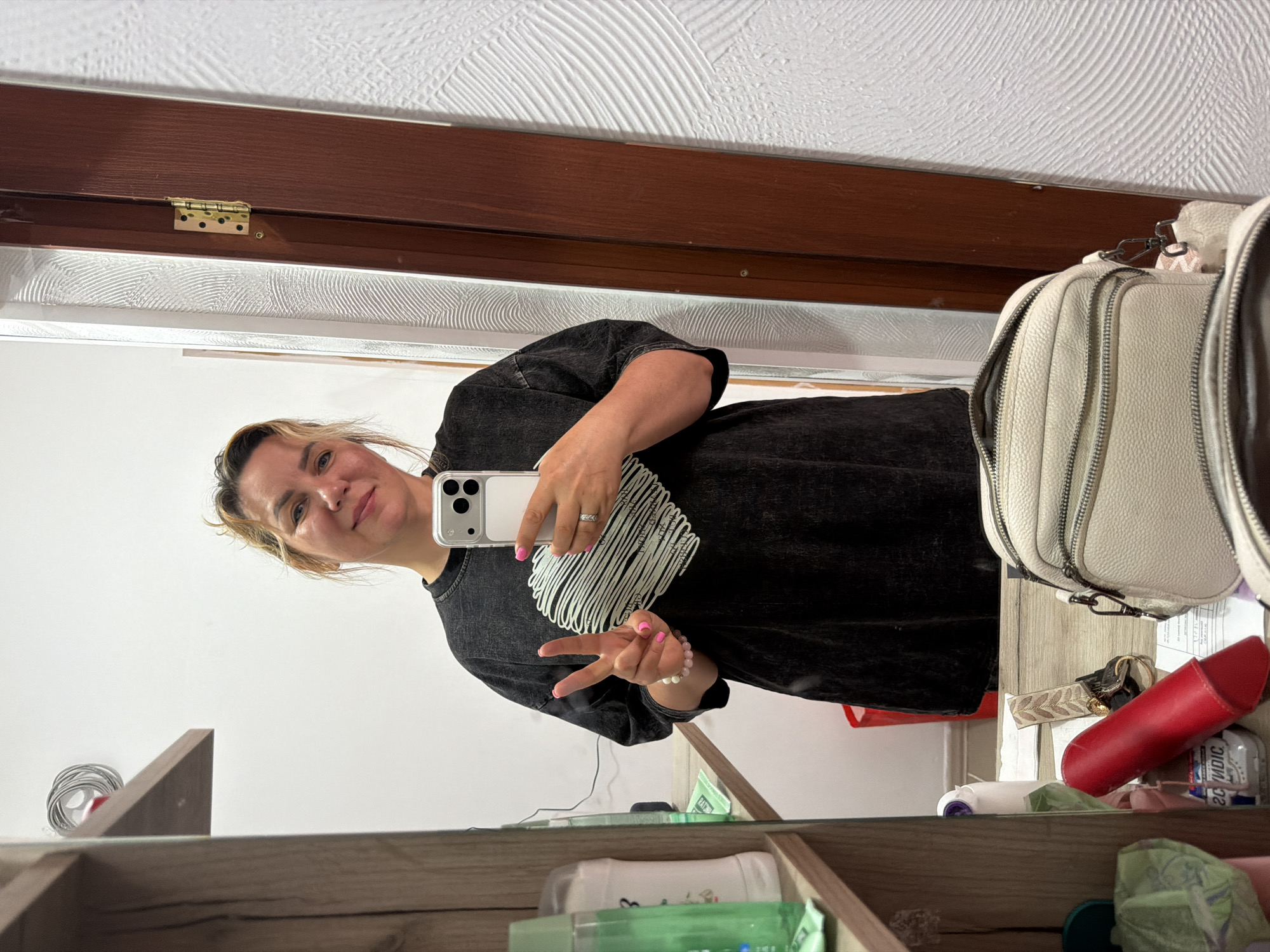 A woman is taking a selfie in front of a mirror. The woman is wearing a dark-colored shirt with a white design, and she is holding a white smartphone. She is making a peace sign with her hand. The reflection in the mirror shows a room with a white wall and a shelf with various objects, including cosmetic products, a red case, and a white purse. The top of the mirror frame is brown wood with brass-colored hinges. The location appears to be indoors, likely a home, but there is no information provided to support that it is located in Leningradskaya, Russia.