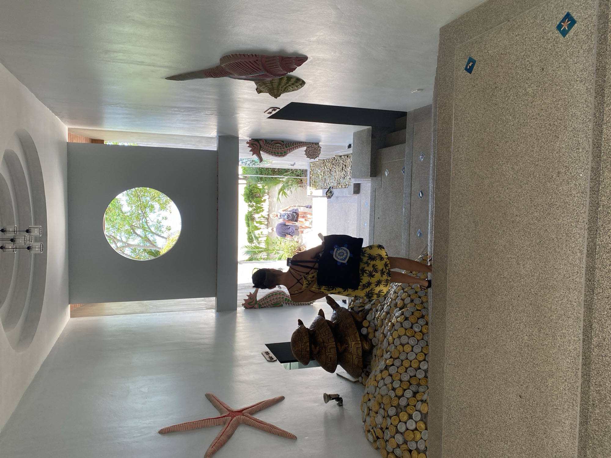 A woman in a yellow floral dress and a black tote bag stands on a staircase, facing away from the camera and looking down a flight of stairs towards an open-air walkway. Two people, one with a backpack, are walking away from the camera on the walkway. The staircase and walkway are decorated with marine-themed art, including a large red starfish on the left wall, a stack of turtle statues on a bed of coins and stones, and a seahorse statue on the right wall. A circular window is visible at the top of the staircase, looking out at trees and greenery. The scene appears to be an entrance or pathway to a resort or hotel in Ko Pha Ngan, Thailand.