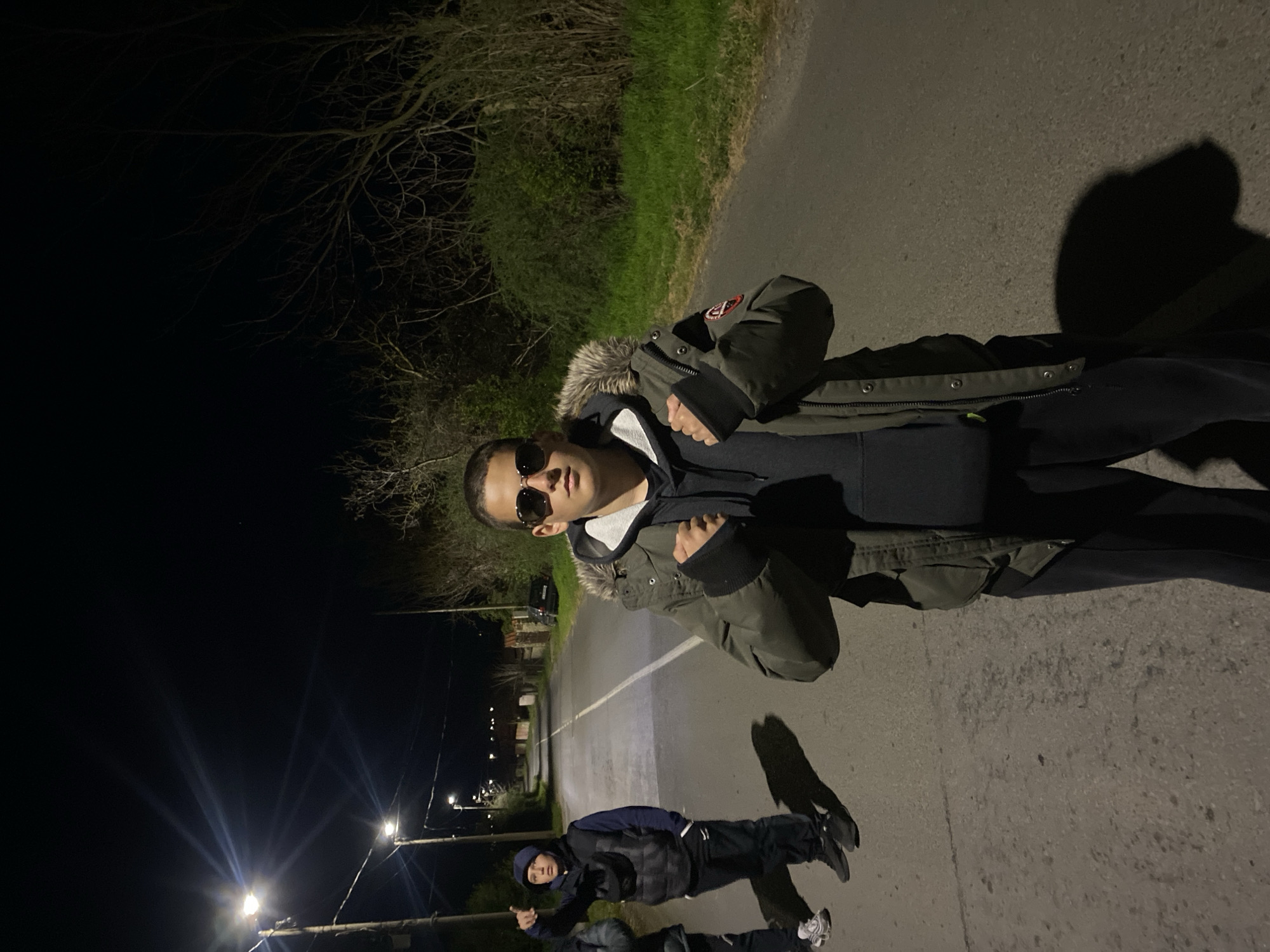 The image shows a nighttime street scene in Goritsa, Bulgaria. Two young males are visible on an asphalt road illuminated by streetlights. One male, wearing sunglasses and a fur-trimmed hooded jacket, stands closer to the camera. A second male, wearing a dark vest and beanie, is visible further down the road. A third male is seen with their head towards the lower right corner of the frame. The surrounding environment includes sparse trees and shrubs along the roadside, and additional streetlights illuminating the scene.
