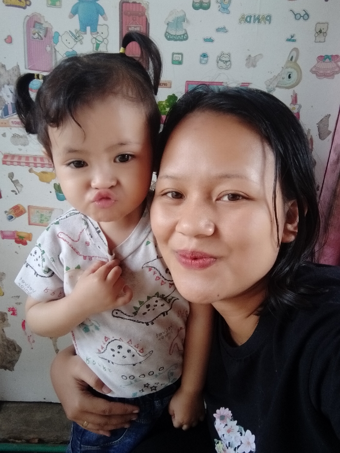 A woman in a black shirt poses for a selfie with a young child with pigtails, wearing a white dinosaur-patterned shirt and jeans. The child is pouting, held by the woman. The background is a patterned wall with various cartoon-like designs. The woman is smiling slightly, and her hair is dark and short.