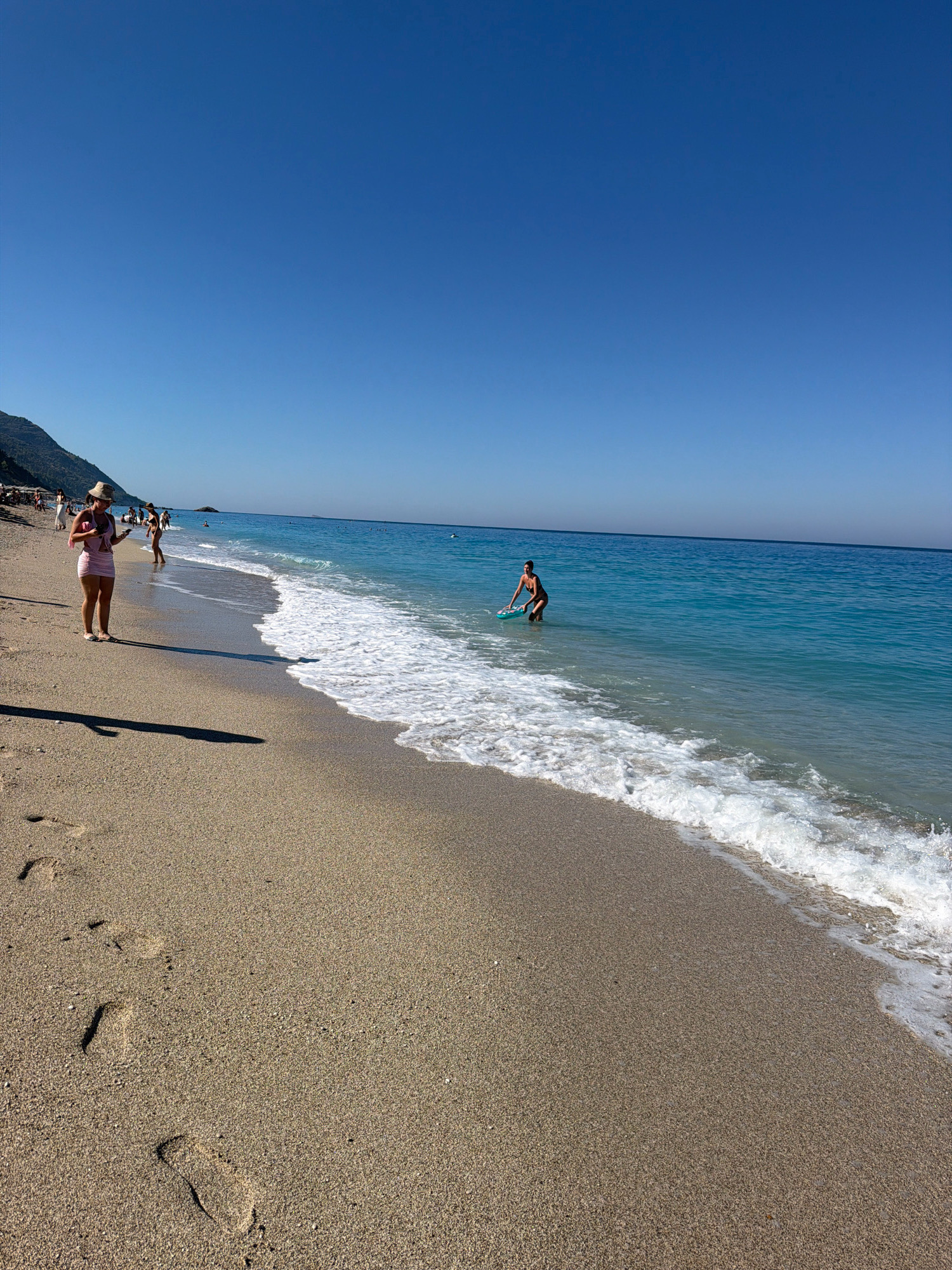 The image depicts a beach scene on the Municipal Unit of Lefkada, Greece. The foreground features a sandy beach with visible footprints leading away from the viewer towards the sea. The sea is a vibrant turquoise blue, with gentle waves lapping at the shore, creating a foamy white edge. In the mid-ground, a person is seen wading in the shallow water, holding a blue inflatable float. Further back, along the shoreline, several other individuals are visible, some walking and others standing. A woman wearing a hat and a pink dress is standing closer to the foreground, looking at her phone. The background shows a clear, bright blue sky and a hilly, tree-covered landscape stretching along the coastline. The overall impression is a sunny day at a popular beach destination.