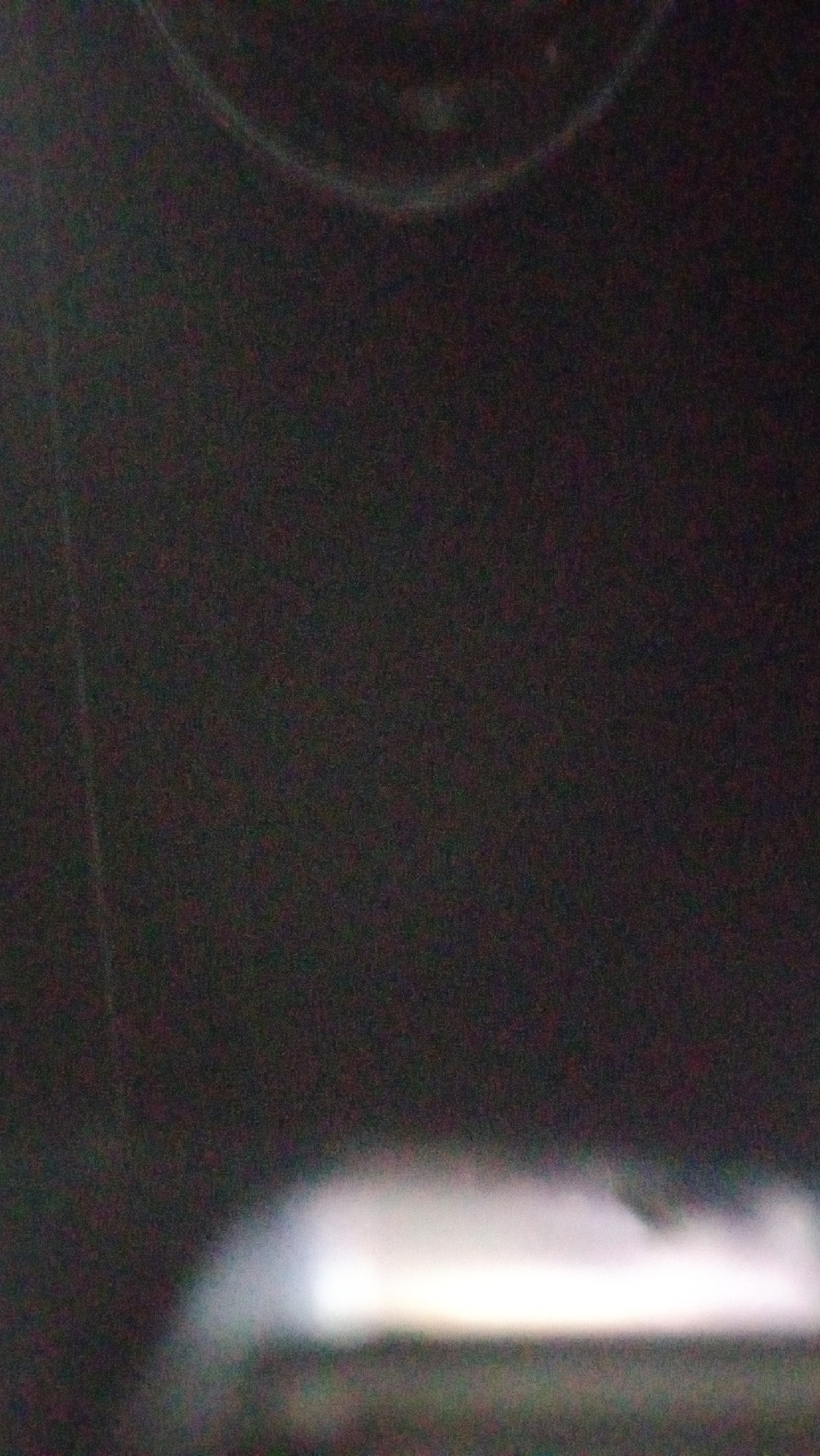 The image is a dark, grainy photograph that appears to have been taken in low light conditions. A bright, horizontal band of light dominates the lower third of the image, suggesting it might be a light source such as a streetlamp or headlights. Above this band, the background is mostly black with some faint, curved lines that could be reflections or the outline of an object. There are no discernible people, specific landmarks, or clear environmental details to place the image in Kuta, Indonesia, or any other specific location. The image quality is poor, making detailed analysis challenging.