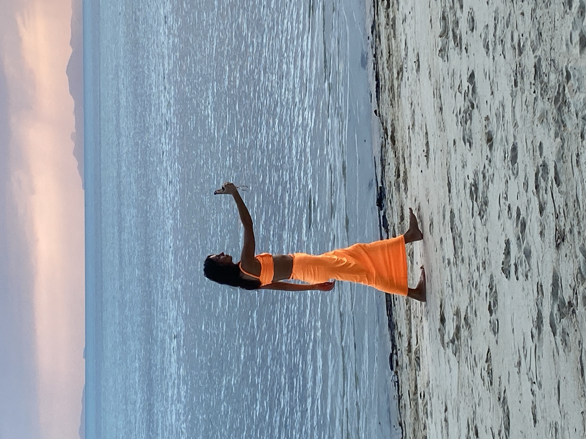 A woman with dark hair, wearing an orange two-piece outfit and a matching long skirt, walks along a sandy beach. She is holding a smartphone in her right hand, appearing to take a selfie or record a video. The ocean is behind her, with calm, blue water reflecting the soft hues of a twilight sky, featuring distant islands or landmasses. The beach sand is light-colored with visible textures and footprints. The overall scene suggests a tranquil moment on the coast of Ko Samui District, Thailand.
