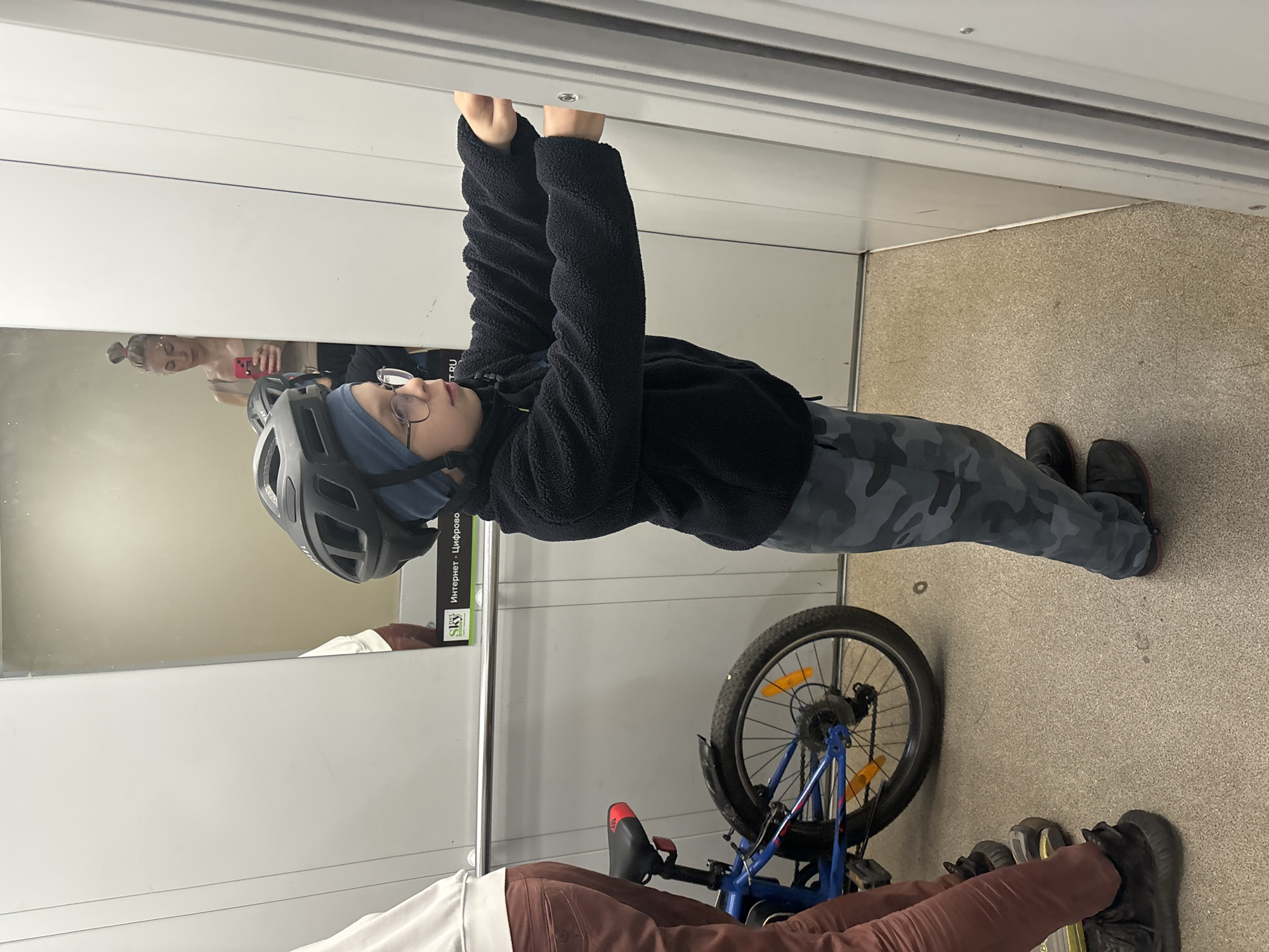 A person in a bicycle helmet and black jacket stands inside what appears to be an elevator, holding onto the top of the doorway. The person is wearing glasses and has a blue head covering. A bicycle is partially visible near their feet, and two other pairs of legs and shoes are also present. A woman's reflection can be seen in a mirror behind the person in the helmet. A sign with Russian text is also visible on the wall. The location is Unknown, Unknown.