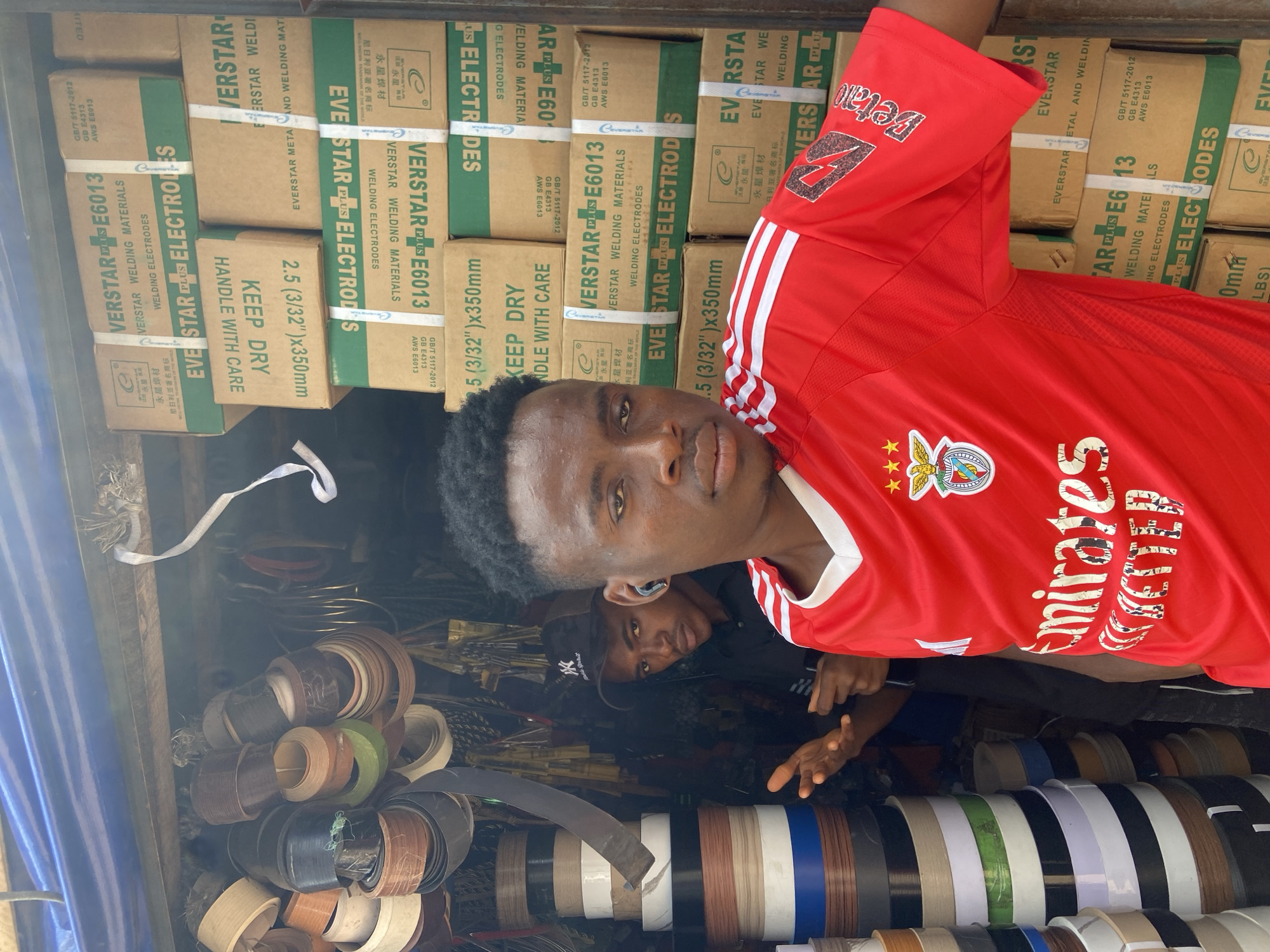 This media file shows a young Black man wearing a red soccer jersey with the "Emirates" logo and a crest on the chest, suggesting a connection to the sport. He is looking at the camera with a relaxed expression. Behind him, another person is partially visible, also looking towards the camera.
The scene appears to be an outdoor market stall, likely in Ogidi, Nigeria, as indicated by the context. The stall is filled with various rolls of material, some resembling edge banding or trim, in different colors and wood-grain patterns. Stacked in the background are several cardboard boxes labeled "VERSTAR E6013 WELDING ELECTRODES." These boxes are in both English and Chinese, indicating the origin and type of product being sold.
The man in the foreground is the primary subject, with the market stall and its wares providing context for the setting. The lighting suggests it is daytime, and the overall atmosphere is casual and perhaps commercial, typical of a market environment. The presence of the welding electrodes and the variety of trim rolls suggests the stall might cater to tradespeople or DIY enthusiasts. The man's gaze and pose are direct and engaging, while the surrounding materials add texture and detail to the scene.