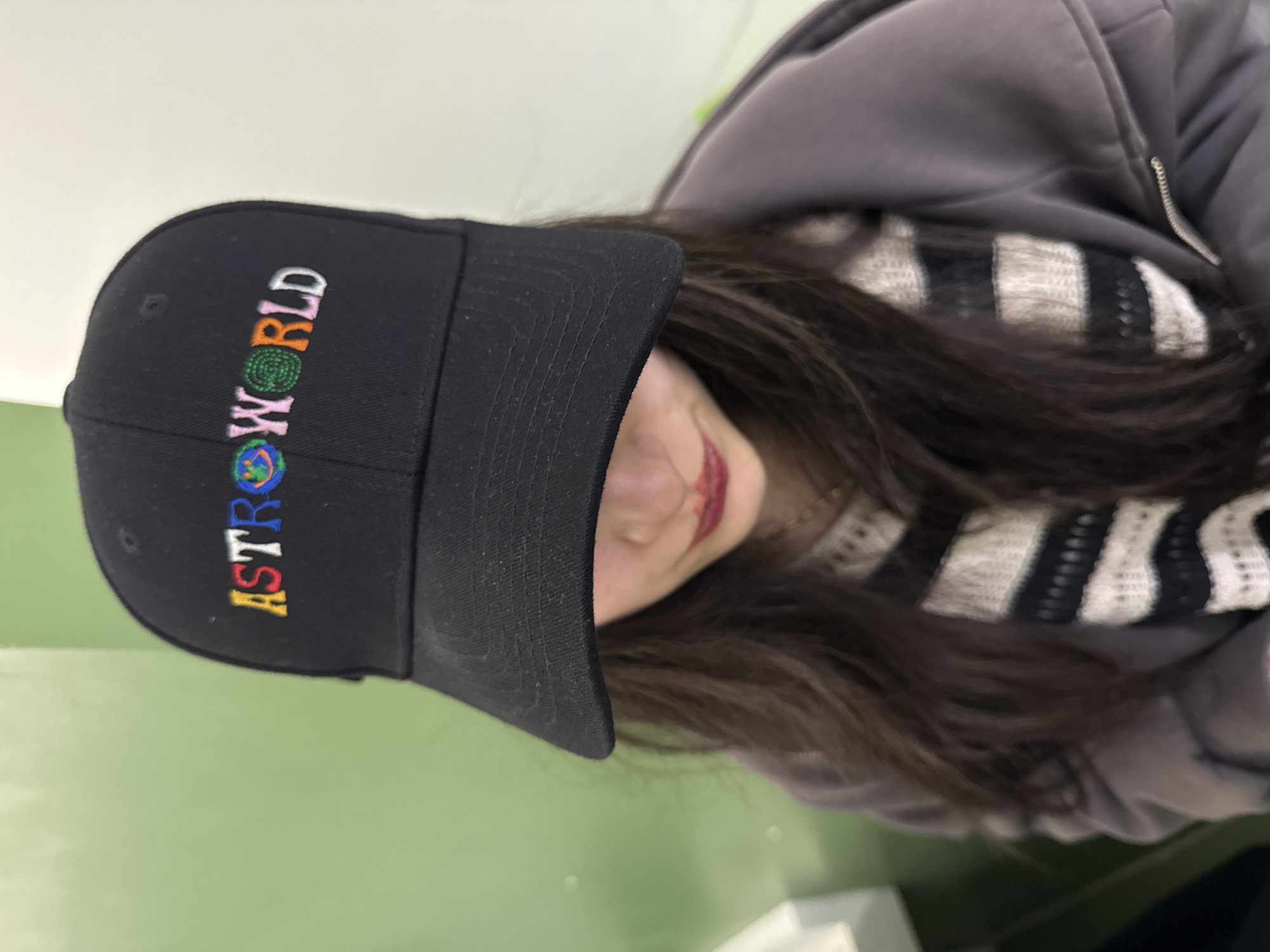 A person is wearing a black baseball cap with "ASTROWORLD" embroidered on it in colorful letters. The person is also wearing a gray zip-up jacket and a black and white striped scarf. The background is a light green wall. There is no indication this photo was taken in Yerevan, Armenia.