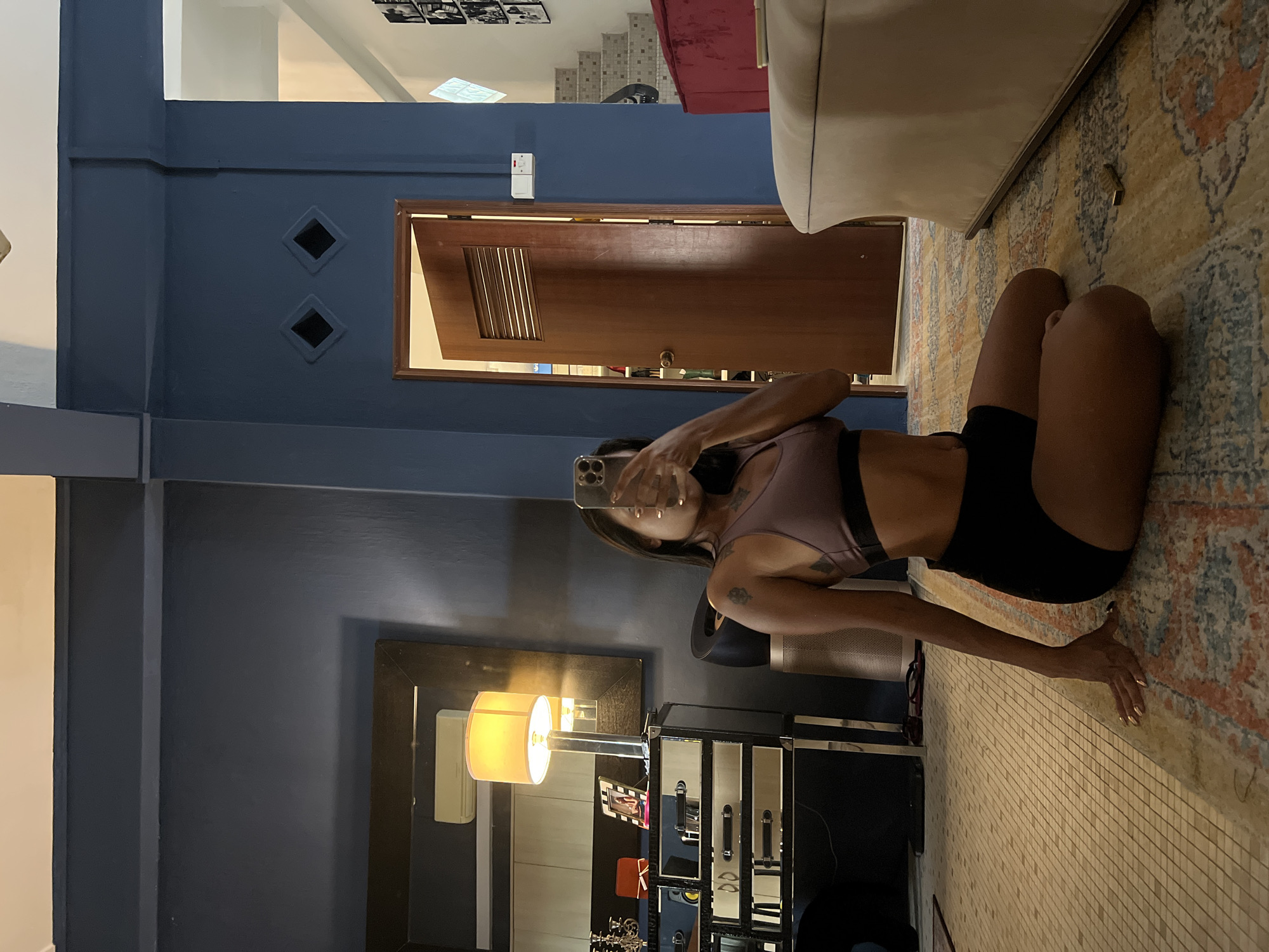 The image depicts an interior scene, likely a home, with a woman in athletic wear sitting on a rug. She is holding a smartphone to take a mirror selfie, obscuring her face. The room features dark blue walls, a large mirror reflecting a lamp and what appears to be an air conditioning unit, and a wooden door with louvered panels. To the left, a dresser with multiple drawers and chrome handles is visible, as well as decorative items on top. The floor is tiled, and a patterned rug is spread across it. In the background, a staircase with patterned risers and a red sofa are partially visible. The overall impression is of a casual, indoor setting. The scene does not contain any specific visual cues that definitively place it in Singapore.