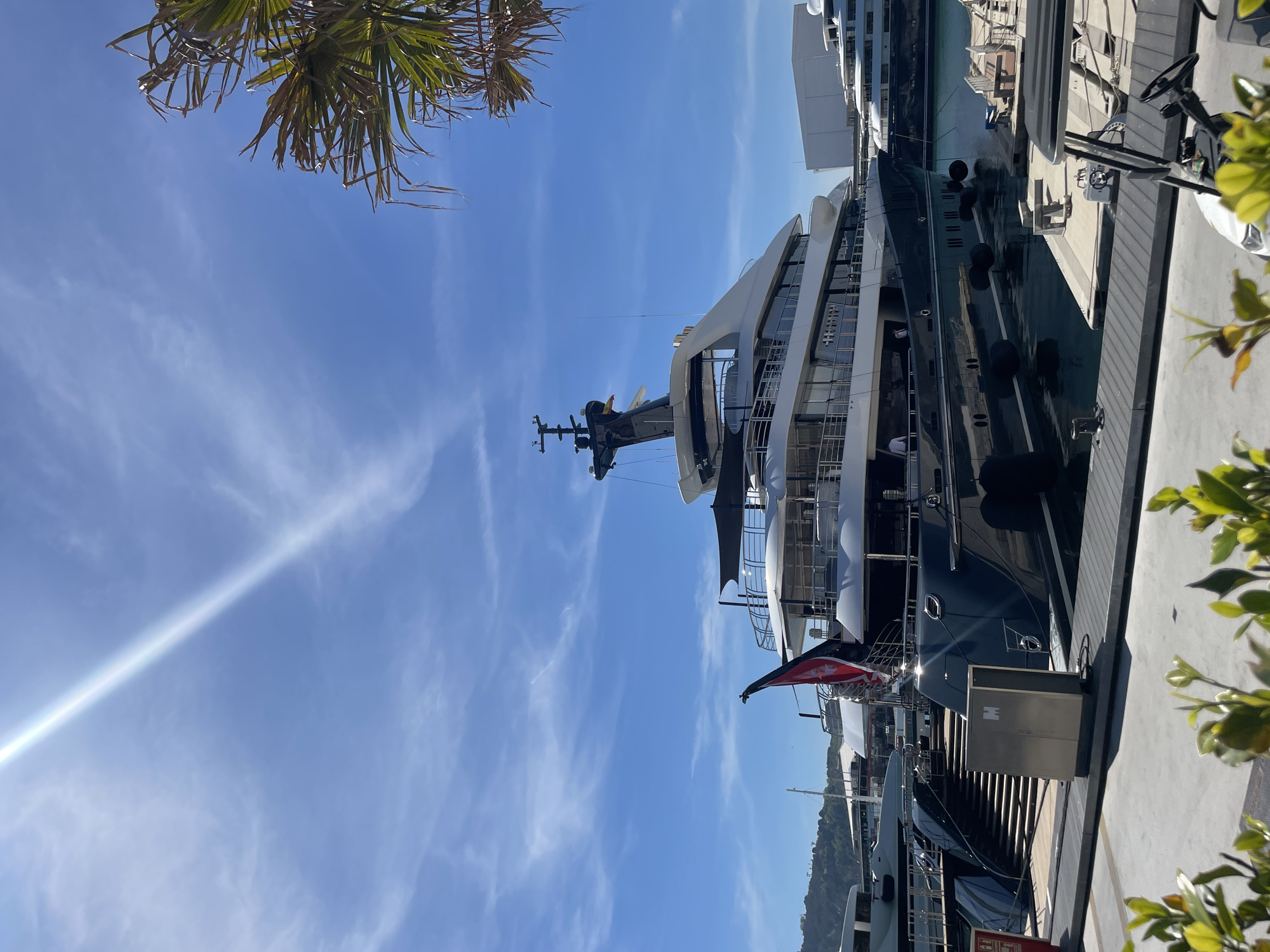 A large superyacht is prominently docked in a sunny marina, dominating the central and right portion of the vertically-oriented photograph. The yacht features a sleek, dark-colored hull and a multi-decked white superstructure with numerous windows and open railing areas. Several black fenders are positioned along its hull where it meets the dock.
The sky is a bright, clear blue with wispy white clouds, and a strong sunbeam or lens flare streaks across the upper left. In the top left corner, the fronds of a palm tree are visible, partially silhouetted. On the opposite side, in the bottom right, green leafy foliage from another plant frames the edge of the scene.
The yacht is moored to a grey paved dock. On the dock, a metal power pedestal is visible with the letter "H" embossed on it. In the background, other large vessels and marina buildings are discernible, along with distant green hills or mountains under the bright light. Calm water in the marina reflects parts of the yacht and the sky. A red and white flag, possibly a courtesy flag, is affixed to the yacht's stern. At the very bottom edge of the image, a red sign with partially visible text appears to read "FIRE". The overall scene depicts a calm, sunny day at a busy and affluent port, consistent with Barcelona, Spain.