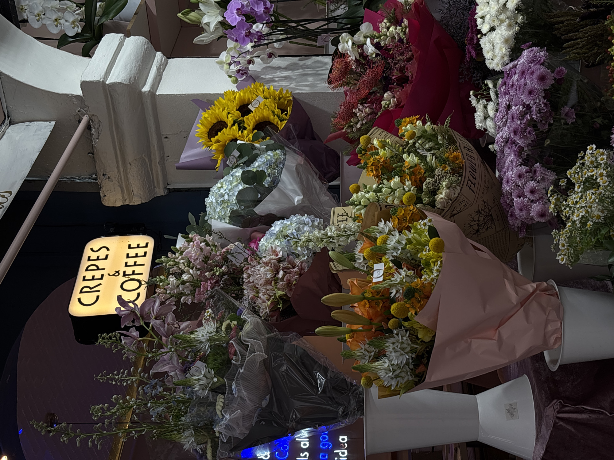 Captured in a shop in Unknown, Unknown, the photograph reveals a display of colorful flower bouquets arranged closely together. The vibrant palette of blossoms features whites, yellows, purples, and oranges. Some of the bouquets are wrapped in materials like brown printed paper and pink or dark plastic.
Illuminated by a sign reading "CREPES & COFFEE," the display's placement near the sign suggests the shop may also sell floral arrangements. The light-up sign has black text on a yellow surface, and is situated on a dark, possibly reflective, surface.
Other elements like a white structure with moldings and a possible building corner hint at the urban setting of the scene. White vases, partially visible, contain some of the bouquets, adding a decorative touch to the overall composition.