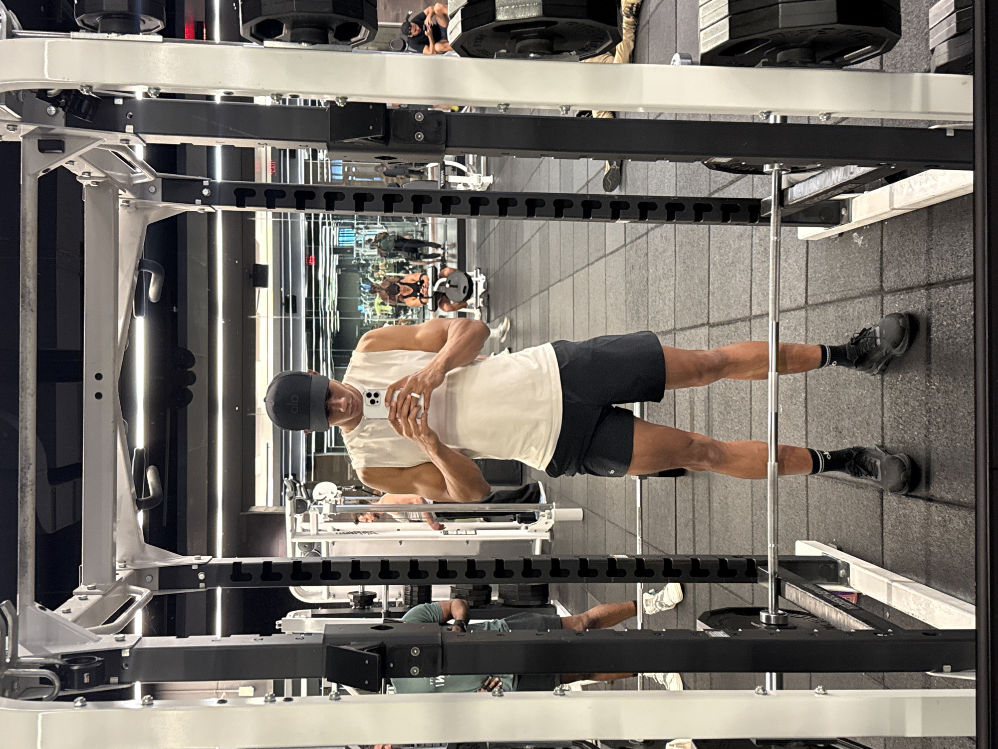 This image, presented sideways, captures a modern fitness center or gym. The setting features dark, square rubber flooring and walls adorned with numerous linear light strips that create a contemporary ambiance and reflect extensively in large wall mirrors.
The primary subject is a fit young man with a tan complexion, positioned within a white and black metal squat or power rack. He is wearing a white sleeveless tank top, black athletic shorts, black socks, and black athletic shoes, topped with a dark baseball cap worn backward that features an "alo" logo. He holds a white smartphone with multiple camera lenses in both hands, seemingly taking a mirror selfie or recording himself. A silver-colored ring is visible on his right ring finger.
Around him, various gym equipment is visible, including stacked weight plates on the rack's holders and a barbell positioned on the floor near his left leg. In the reflections of the large mirrors behind him, other gym-goers are discernable. One individual in a dark top is seen seated, while another wearing a light green t-shirt appears to be preparing for exercise or resting. The gym appears well-maintained and brightly lit, suggesting an indoor daytime setting.
