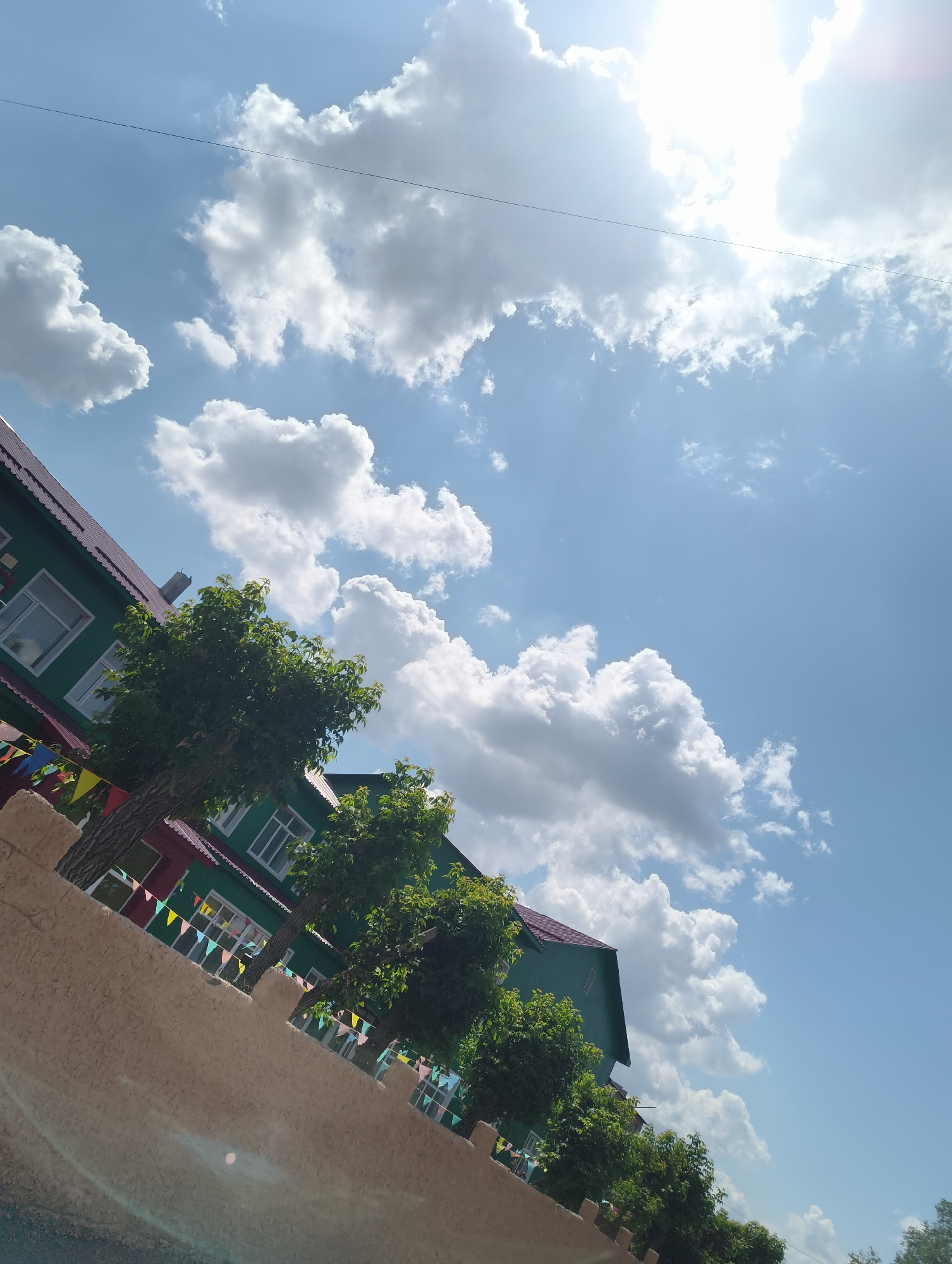 An eye-level shot captures a bright, sunny day with a blue sky dotted with white, fluffy clouds. The sun is a strong, white presence in the upper right corner, with light rays emanating outward. In the foreground, a textured, beige wall runs diagonally across the bottom of the frame. Behind the wall, a green building with a maroon roof is visible, featuring multiple windows. A string of colorful triangular flags hangs along the building's facade. Two trees with lush green foliage are positioned in front of the building. A thin, dark wire is visible horizontally across the upper portion of the sky.