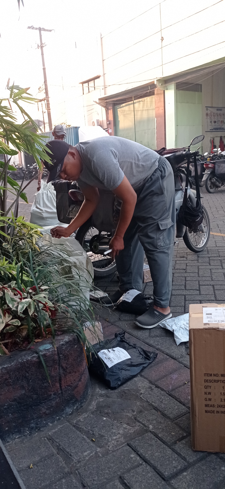 In Jemundo, Indonesia, a man wearing a grey t-shirt and grey cargo pants bends over to attend to a motorcycle. He is wearing a black baseball cap and black slip-on shoes. Next to him, on the brick-paved ground, are several black plastic packages, a white plastic bag, and a cardboard box. The box has text printed on it, including "ITEM NO: MI", "QTY : 1", "N.W : 1.5", "G.W : 2.1", and "MADE IN IND". In the background, a second man in a grey t-shirt and grey cap stands near a large white bag, and several motorcycles are parked. Lush green plants with variegated leaves are in the foreground, planted in a dark brown planter. The scene appears to be an outdoor area, possibly a loading dock or a street-side location.