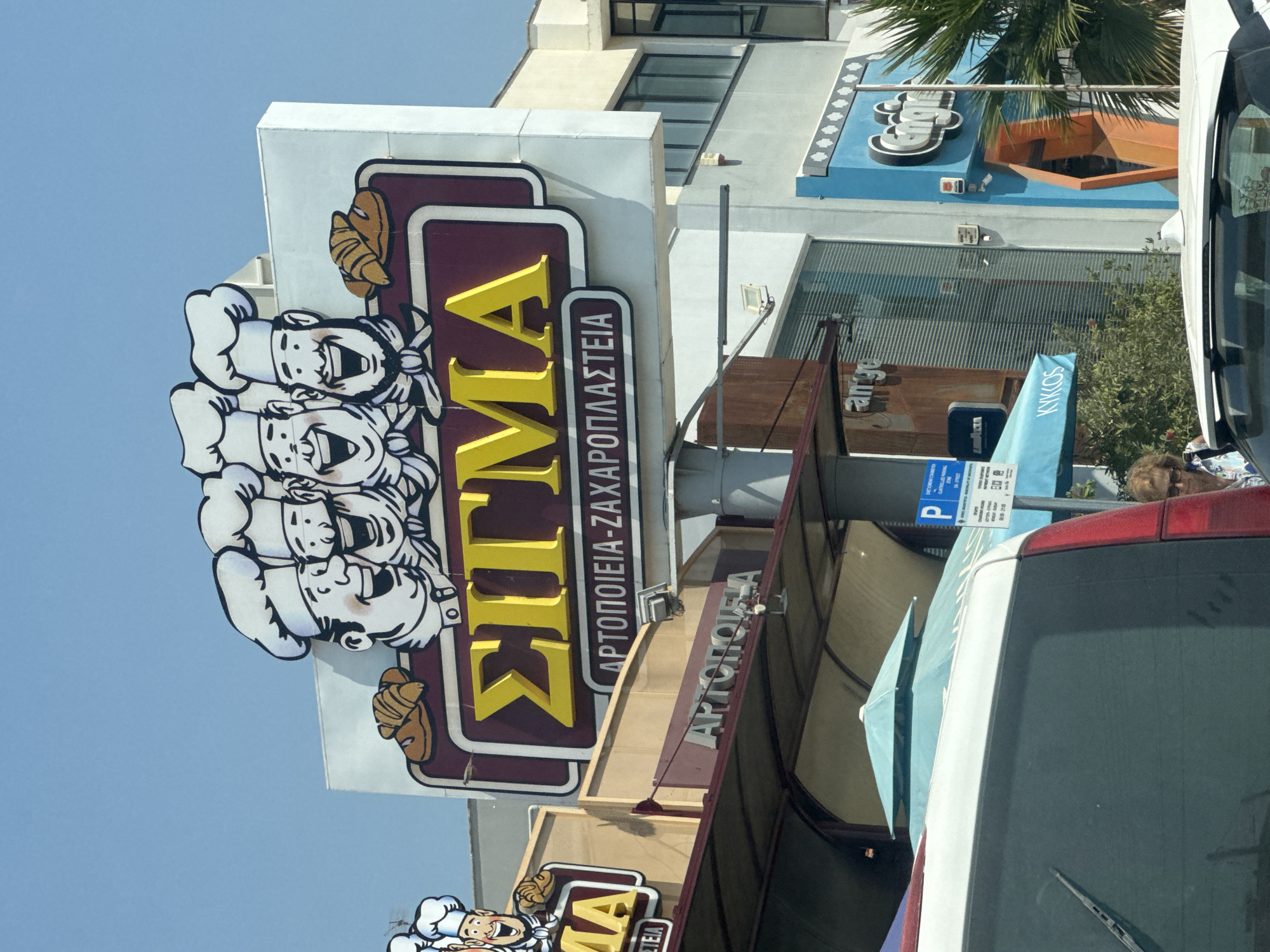 The image shows a bright, sunny day in Agios Athanasios Municipality, Cyprus. A large, colorful sign for "ΣΙΓΜΑ" bakery dominates the upper portion of the frame. The sign features cartoon chefs and loaves of bread. Below the bakery sign, the words "ΑΡΤΟΠΟΙΕΙΑ-ΖΑΧΑΡΟΠΛΑΣΤΕΙΑ" (Bakery-Confectionery) are visible.
In the foreground, the rear of a white vehicle is captured, with a partial view of a woman with glasses and light brown hair looking to her right. To the right of the vehicle, a blue parking sign is mounted on a pole. In the background, various shops and buildings are visible, including a store with a blue facade and an orange entrance. Palm trees and a light blue umbrella add to the Mediterranean atmosphere. The overall scene appears to be a commercial area with shops and businesses.