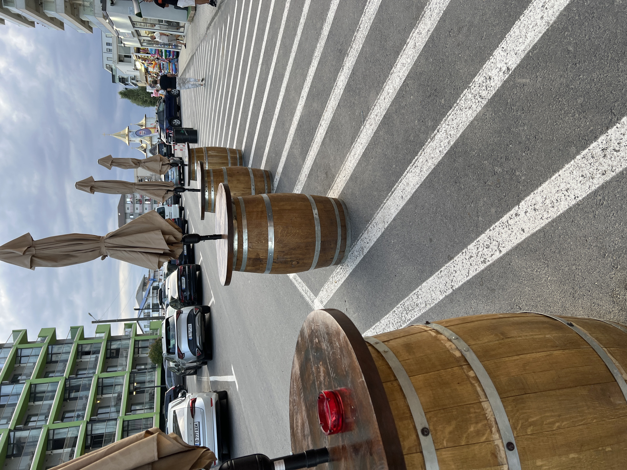 The image depicts an outdoor scene in Năvodari, Romania, likely along a boardwalk or promenade, featuring a row of wooden barrel tables with folded umbrellas, set up on a paved area with white diagonal stripes. In the background, a modern green-facade building stands to the left, while to the right, a street lined with shops and people is visible, leading towards a building with a golden dome. Several cars are parked on the street. A red ashtray is placed on the table in the foreground. The sky is overcast.