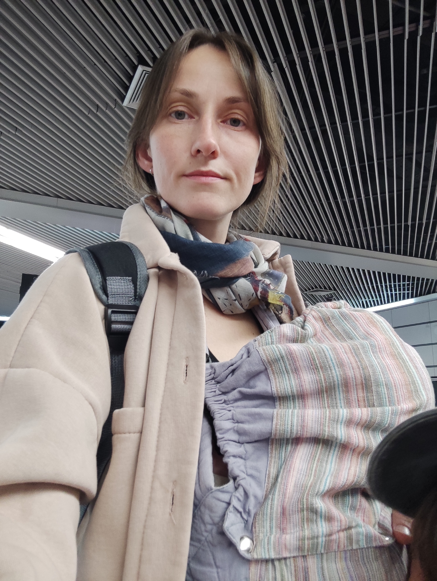 An adult female is depicted in a chest-up, eye-level perspective, centrally positioned and looking directly into the camera. She possesses light brown hair, fair skin, and light-colored eyes, with a neutral facial expression. She is dressed in a light beige or pale pink jacket worn over a dark garment, and a multi-patterned scarf, predominantly blue and grey with accents of other muted colors, is tied around her neck. A black and grey padded strap, consistent with a backpack, is visible over her left shoulder (viewer's right). On her right side (viewer's left), a baby carrier or sling is visible, partially obscuring an infant whose dark head or hat is discernible in the lower right corner of the frame. The carrier features a section with vertical pastel stripes (pink, green, blue, white) adjacent to a section of solid greyish-purple fabric. The background reveals an indoor environment with a distinctive ceiling structure comprising numerous parallel, light-colored horizontal slats or louvers against a dark overhead void, characteristic of a modern public building such as an airport terminal in the city of Sochi, Russia. A white rectangular ventilation grate is visible in the upper right portion of the background. Illumination appears to originate from overhead sources within the ceiling structure.