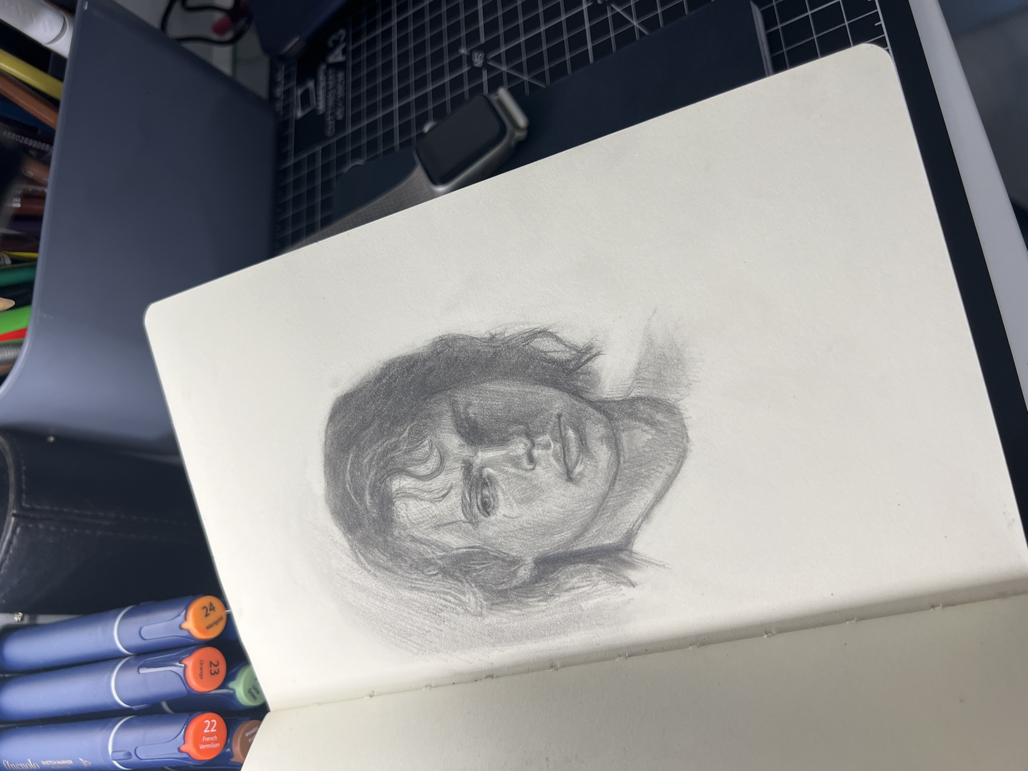 A close-up, high-angle shot captures a pencil sketch of a young man's face within an open sketchbook. The sketch, rendered in grayscale, features the subject with wavy, shoulder-length hair and a serious expression. Light and shadow are used to define his features, creating a sense of depth and volume. The sketchbook page is textured and slightly worn at the spine.
In the background, to the left of the sketchbook, are three blue markers with orange caps, labeled with numbers 22, 23, and 24 and descriptive words. Behind the markers, a dark, leather-bound object is visible. Further back, a gray bin filled with colored pencils and pens stands out. To the right of the sketchbook, a black cutting mat with a grid pattern is partially visible, with a silver smartwatch with a black band resting on it. The overall scene suggests an artist's workspace.
