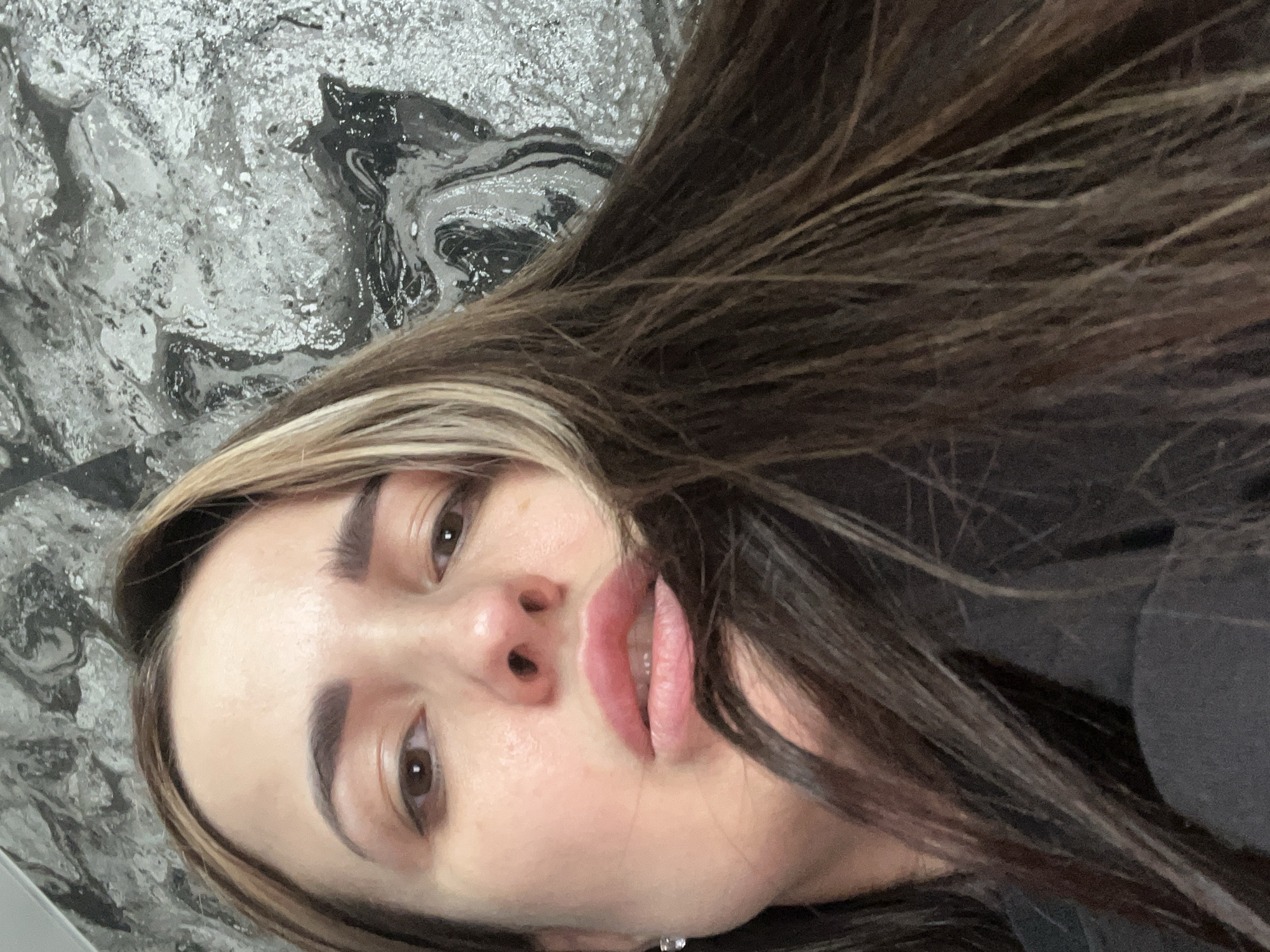 A close-up, slightly off-center portrait of a woman with brown hair and blonde highlights. The background appears to be a textured wall with a marbled pattern in shades of grey, silver, and black. No location is determined or named as city. Unknown.
