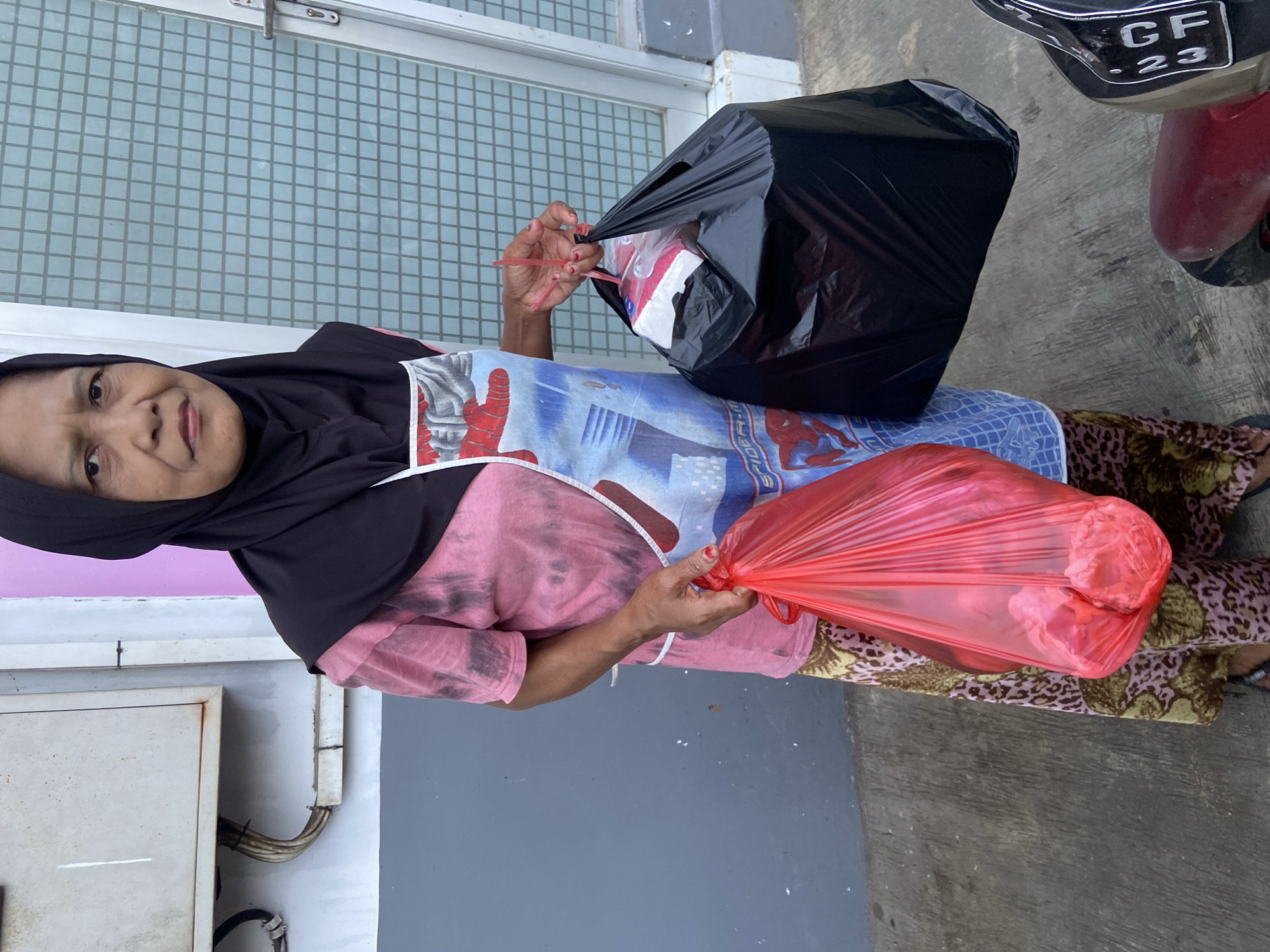 In Jember, Indonesia, an elderly woman wearing a black hijab, a pink and black tie-dye shirt, an apron with a superhero pattern, and patterned pants stands on a concrete surface in front of a light blue tiled wall and a gray wall. She is holding a large black plastic bag in her right hand and a red plastic bag in her left hand. A motorcycle with a license plate that includes "GF 23" is visible on the right side of the frame. An electrical box with exposed wiring is on the left side of the frame.