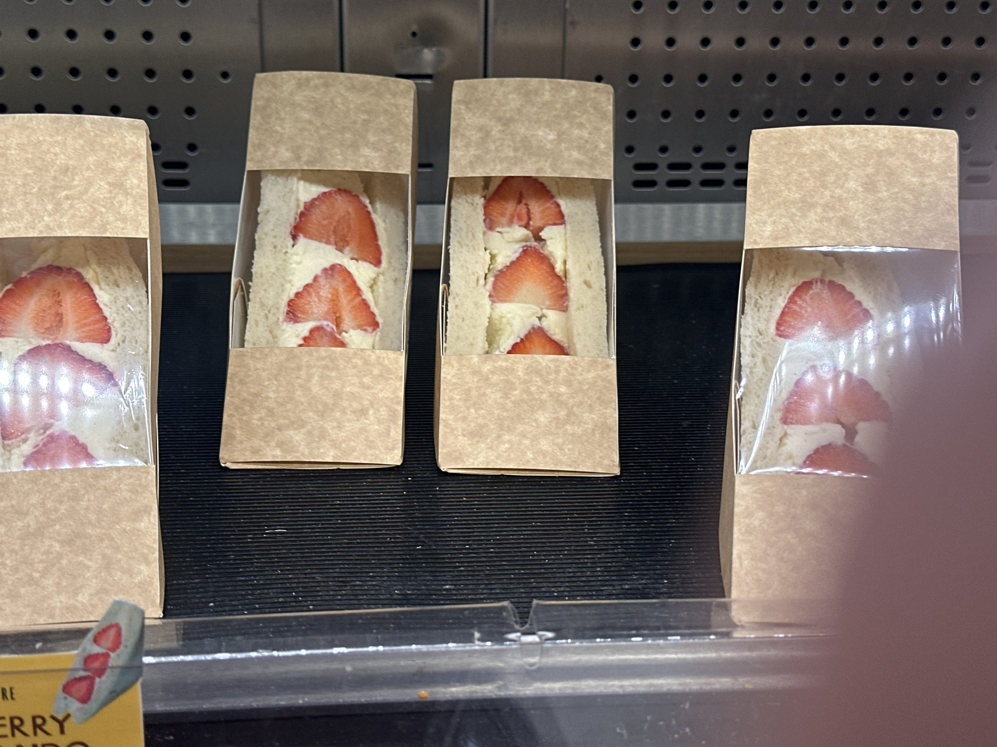 In a well-lit display case in London, United Kingdom, four packages of strawberry sandwiches are arranged in a row on a black surface. The sandwiches consist of white bread, cream, and sliced strawberries, and each package is wrapped in clear plastic and packaged in a small brown paper box. The background is a metallic surface with evenly spaced holes. A small portion of a label with a strawberry design is visible at the bottom left. A hand is partially visible on the right side.
