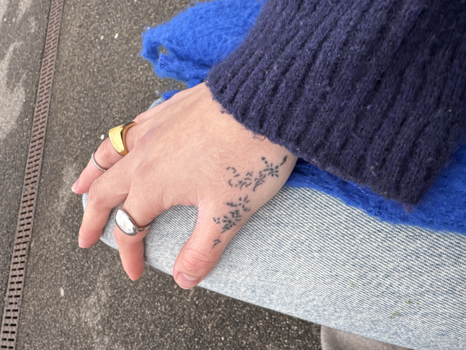 A person's hand with a floral tattoo, adorned with three rings, rests on their denim-clad thigh. The hand is positioned between the person's dark blue sweater sleeve and a vibrant blue, possibly knitted, garment. The rings include a gold square ring, a silver ring with small indentations, and a silver signet ring. The scene is captured outdoors, with a section of pavement visible in the background.