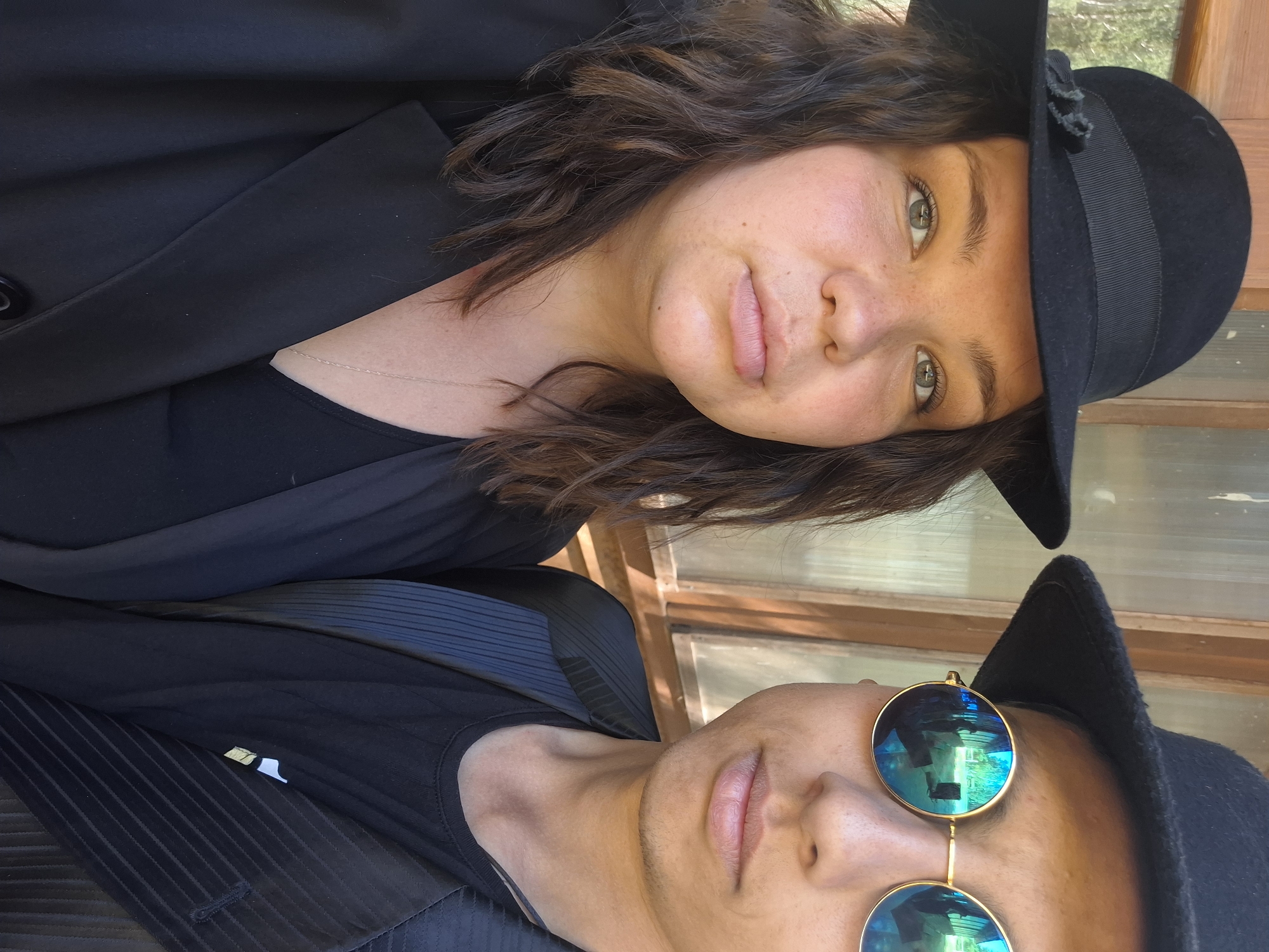Two people are posing for a photograph. The person on the top of the image is a woman with brown curly hair, wearing a black fedora and a black blazer. She has fair skin and light-colored eyes. The person on the bottom of the image is wearing blue mirrored round sunglasses and a black fedora. They are wearing a black shirt and a black pinstriped vest. The location is difficult to discern, but there is a window with a wooden frame in the background. The photograph does not provide enough information to place the location in Chaikovsky, Russia.