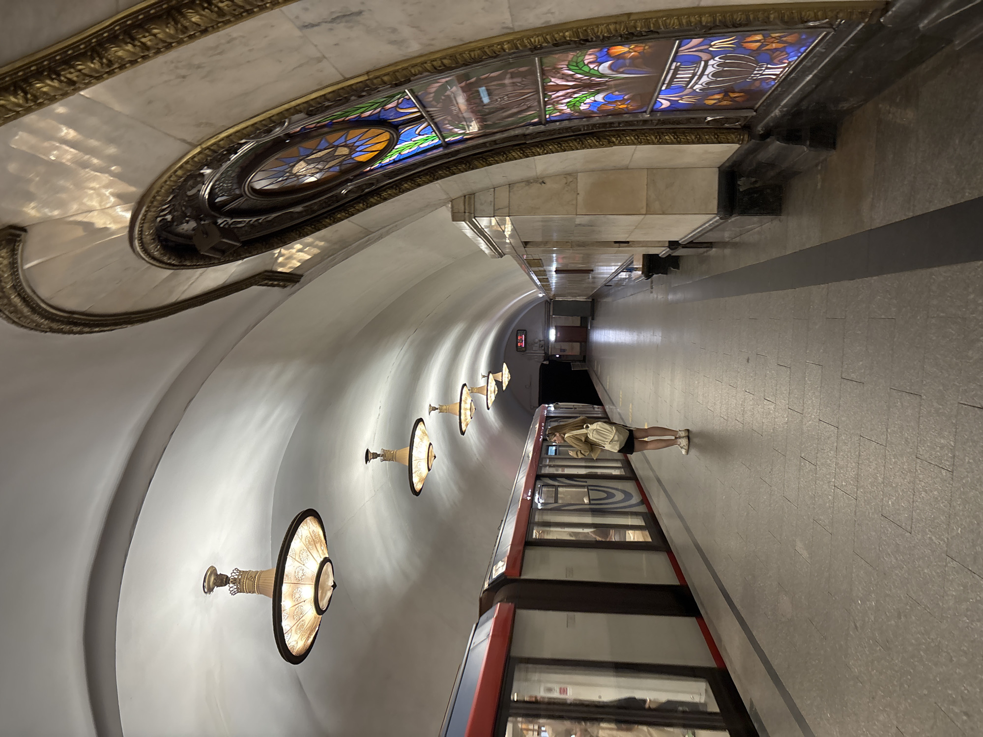 The image captures a scene within a Moscow, Russia metro station. The architecture features a vaulted ceiling adorned with ornate lighting fixtures and decorative stained-glass panels. A metro train is positioned at the platform, and a person stands on the platform next to the train. The platform is tiled.