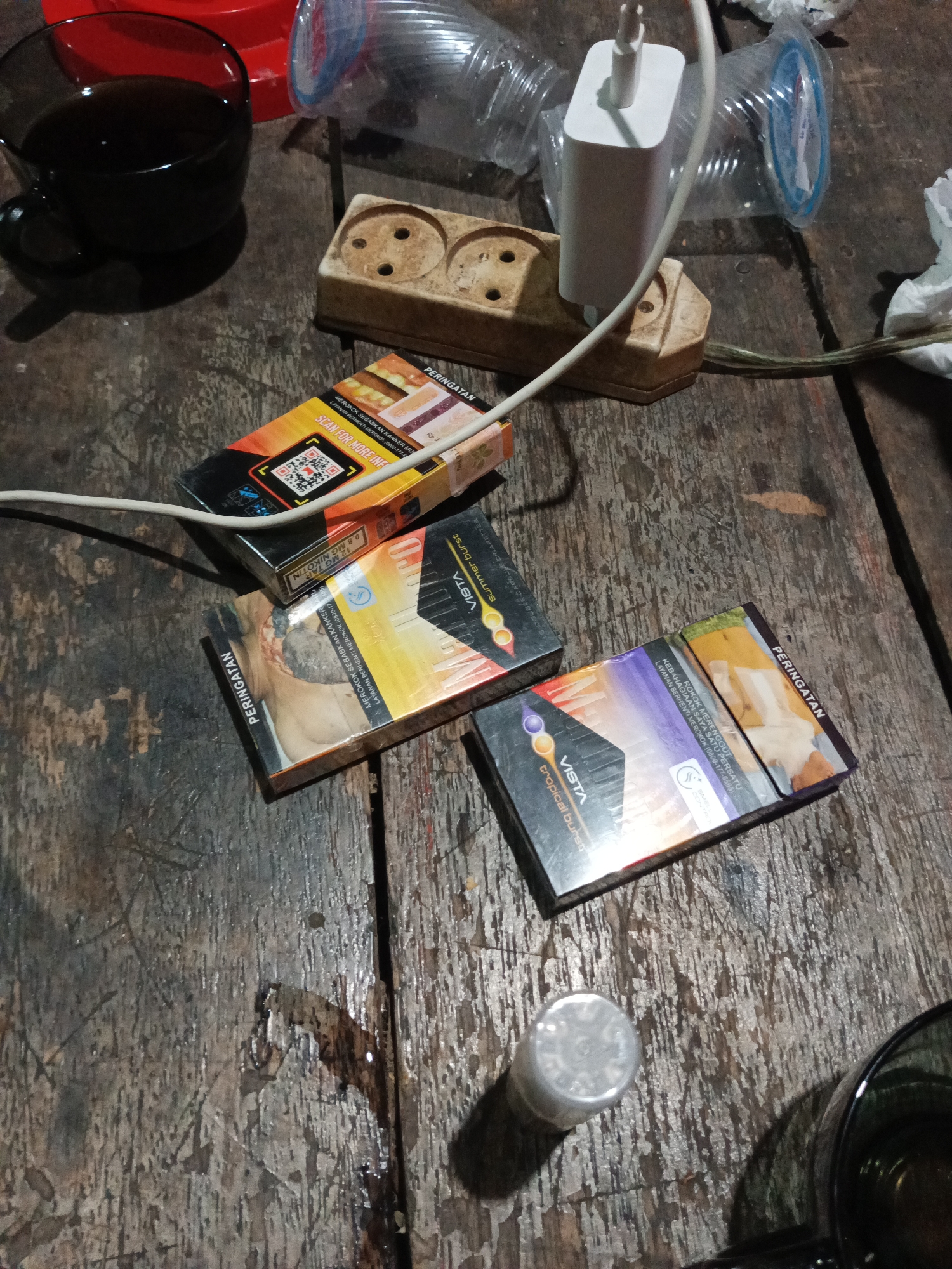 The image depicts a dimly lit wooden table surface. On the table, there are several items, including a dark-colored mug containing a dark liquid, a beige electrical outlet strip with a white power adapter plugged into it, two packs of "VISTA" brand cigarettes (one orange and yellow, the other purple and yellow), and a clear plastic bottle. A white charging cable is draped across the outlet strip and extends towards the cigarette packs. There are also some crumpled white paper items visible. The wooden surface appears worn and somewhat damp in places, with visible grain and texture. The overall setting suggests a casual, possibly indoor, environment.