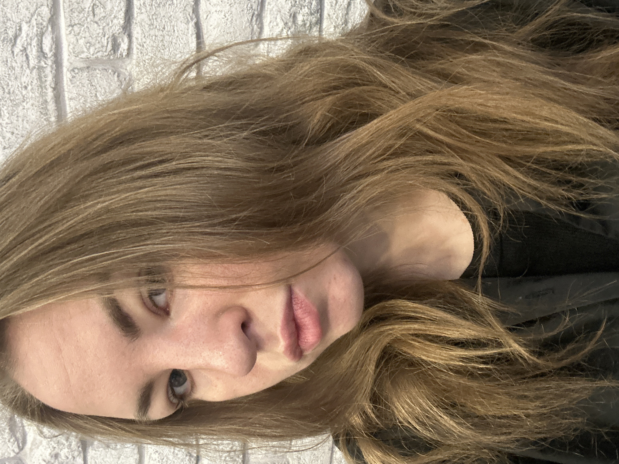 A close-up, slightly off-center portrait shot captures a female subject, estimated to be a young adult, from the mid-chest upward. Her head is angled slightly, and her light-colored eyes are directed towards the camera with a direct gaze. Her long, light brown hair, styled with loose waves, frames her face and falls over her shoulders, partially obscuring the sides of her face and neck. Visible forehead lines are present. Her lips are closed, displaying a natural pinkish tone with minor imperfections on the lower lip. She is dressed in a dark, possibly black, outer garment over a black inner layer. The background consists of a textured, light-colored brick or stone wall with visible mortar lines. The location is in the city of Unknown, Unknown.
