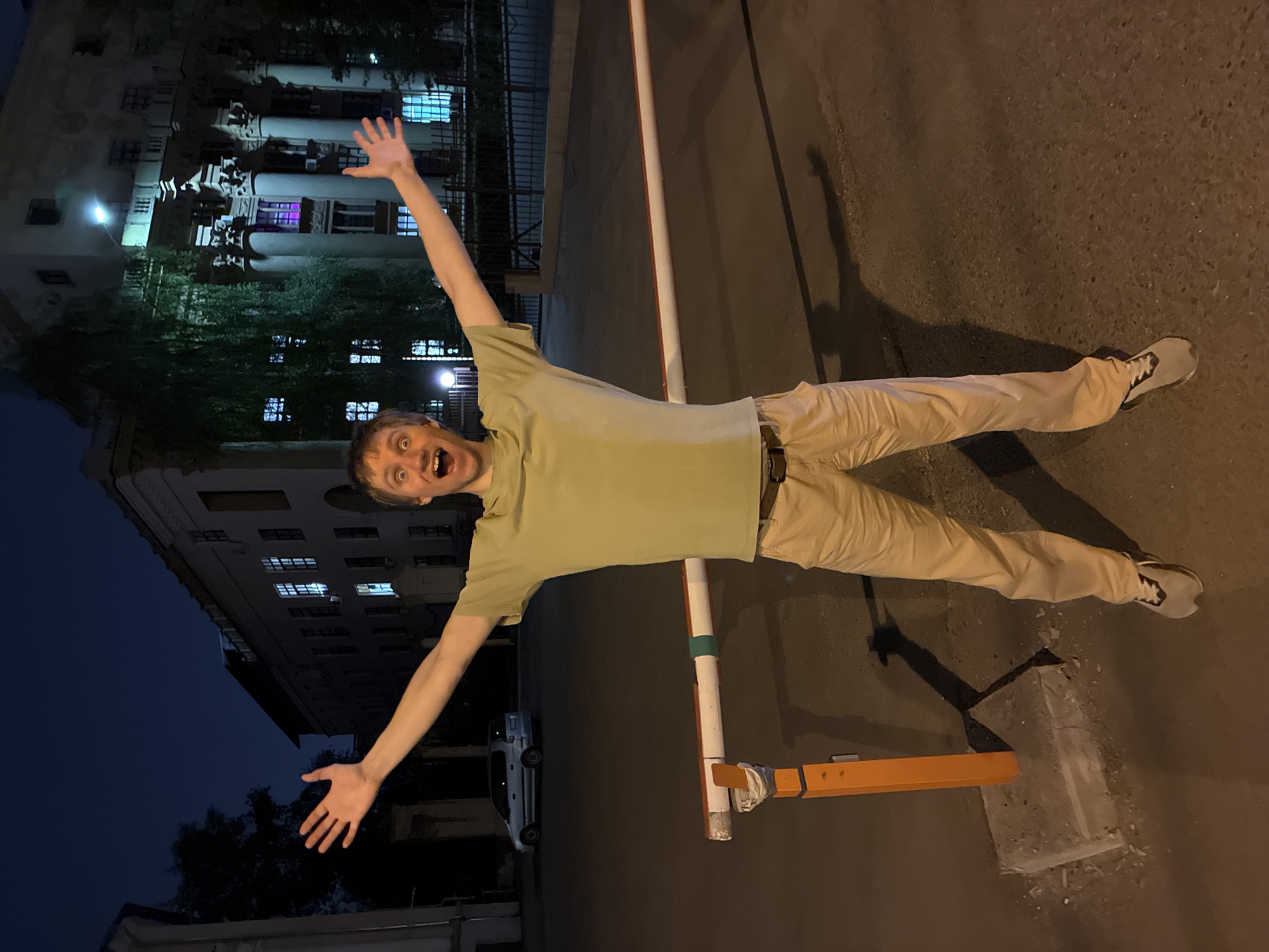 A man stands in front of a building at night, arms outstretched. The building has lit windows and is likely located in Moscow, Russia. The man is wearing a light green t-shirt, khaki pants, and gray sneakers. He is positioned next to a metal barrier painted white and orange. A white car is partially visible in the background, parked near the building. The area is illuminated by streetlights, casting shadows on the asphalt.