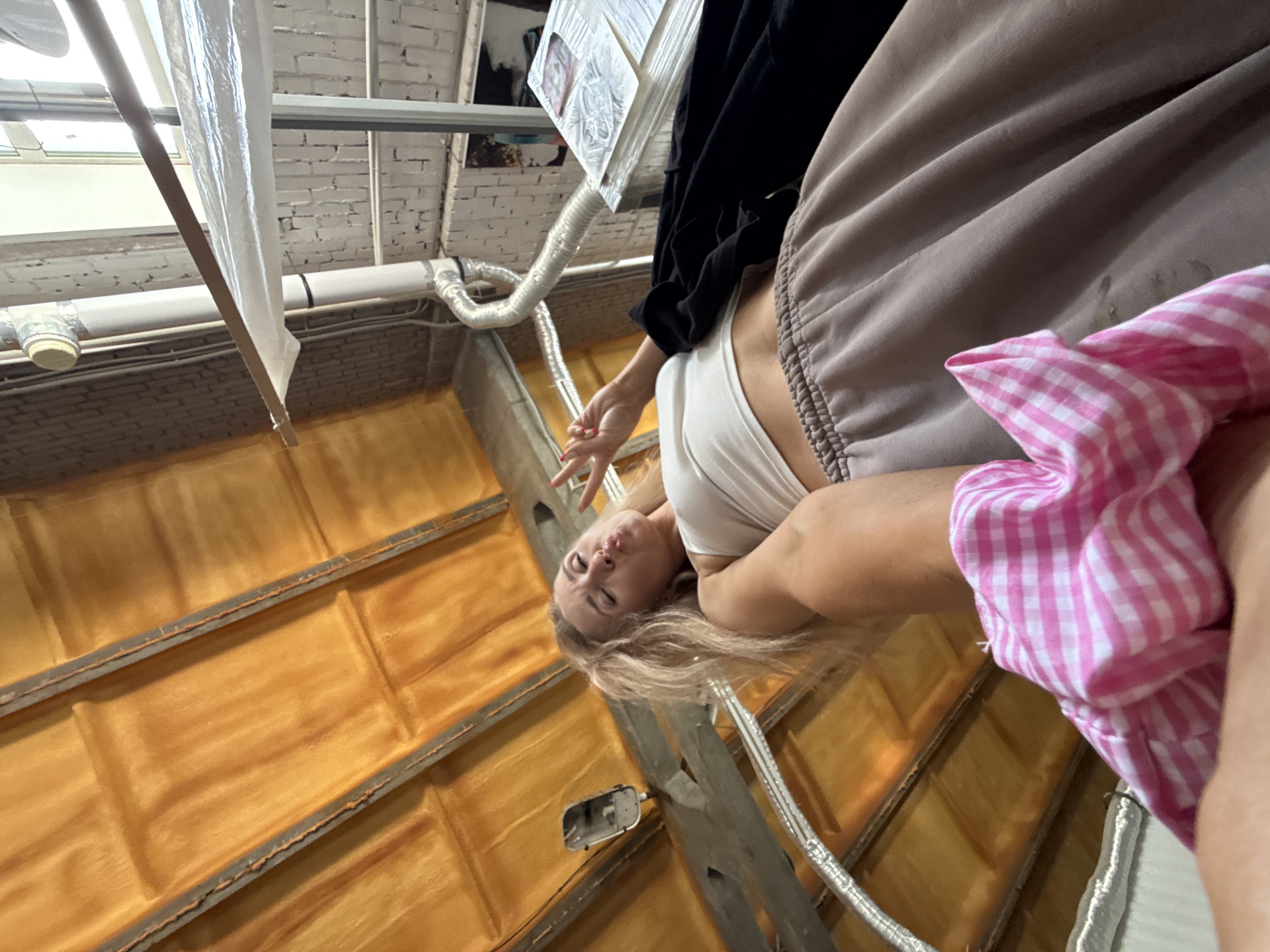 The image, taken in the city of Chaikovsky, Russia, shows a woman lying down, positioned at an unusual angle relative to the camera, creating a disorienting perspective. She is wearing a white top and a light brown garment, with a pink and white checkered item partially obscuring the view. The background reveals elements of what appears to be an industrial or unfinished interior space, with orange-colored, possibly insulated, surfaces. Metal framing and ventilation pipes are visible along with white brick ceiling and a hanging light. The overall impression is that the woman is resting in a construction or storage-like environment.