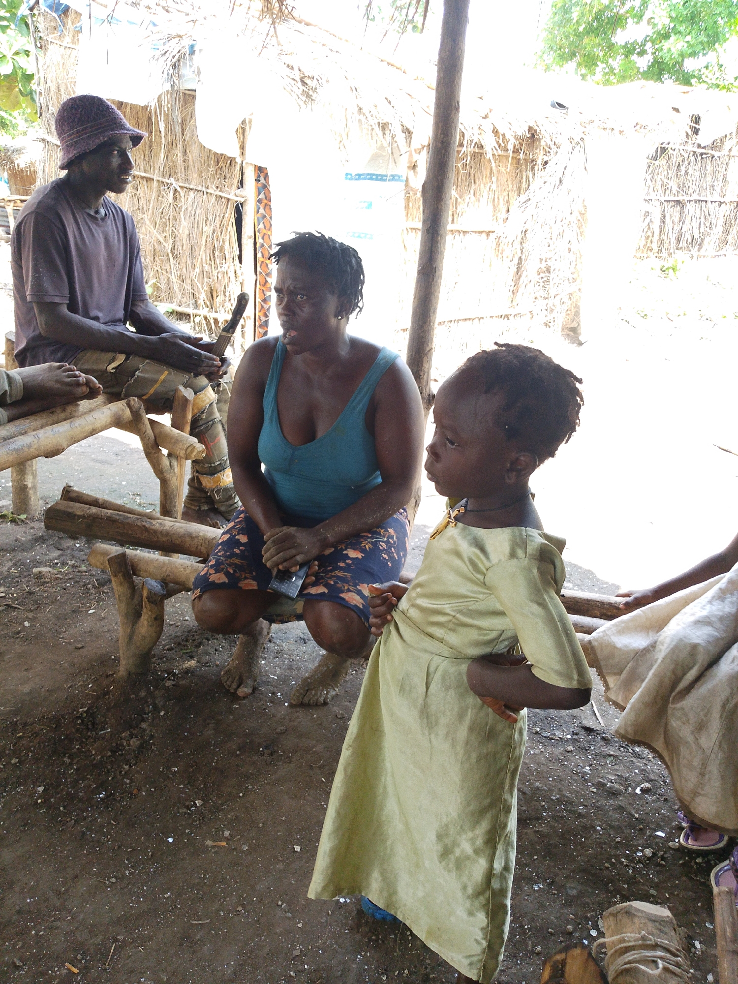 In Otulu, Nigeria, three people are gathered under a thatched roof structure. A man wearing a purple bucket hat and a t-shirt sits on a wooden bench, holding a knife. To his right, a woman in a blue tank top and a floral skirt is seated, holding a mobile phone. A young child in a light green dress stands beside the woman, looking to the side. The ground is covered in dirt and debris, with a wooden structure and more thatched roofing visible in the background. The overall scene depicts a casual gathering in an outdoor, rural setting.