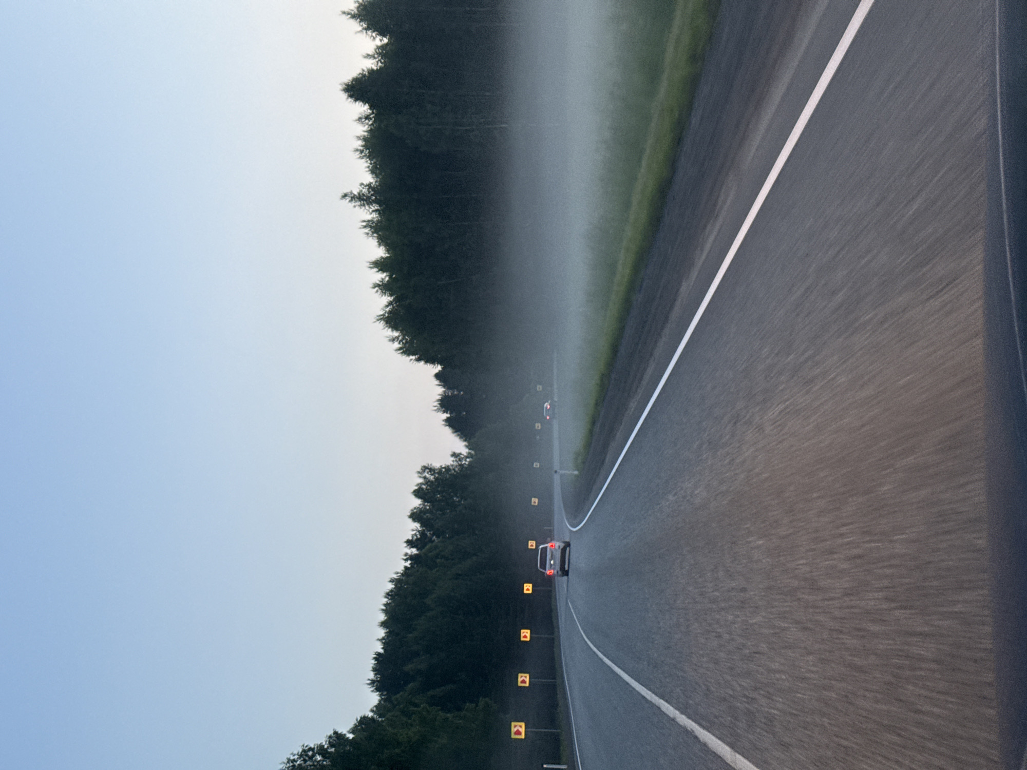 The image depicts a view from a moving vehicle on a highway in Sosnovo, Russia, as indicated by contextual information. The highway curves to the right, with a light fog or mist present in the grassy area alongside the road and extending towards the dense coniferous forest lining the left side. Several yellow, diamond-shaped directional signs are visible along the left side of the road, indicating curves. A silver car is seen driving ahead on the highway, with its brake lights illuminated. The sky is clear and light blue, suggesting either early morning or late evening light conditions. The road surface shows motion blur, consistent with a vehicle in motion.